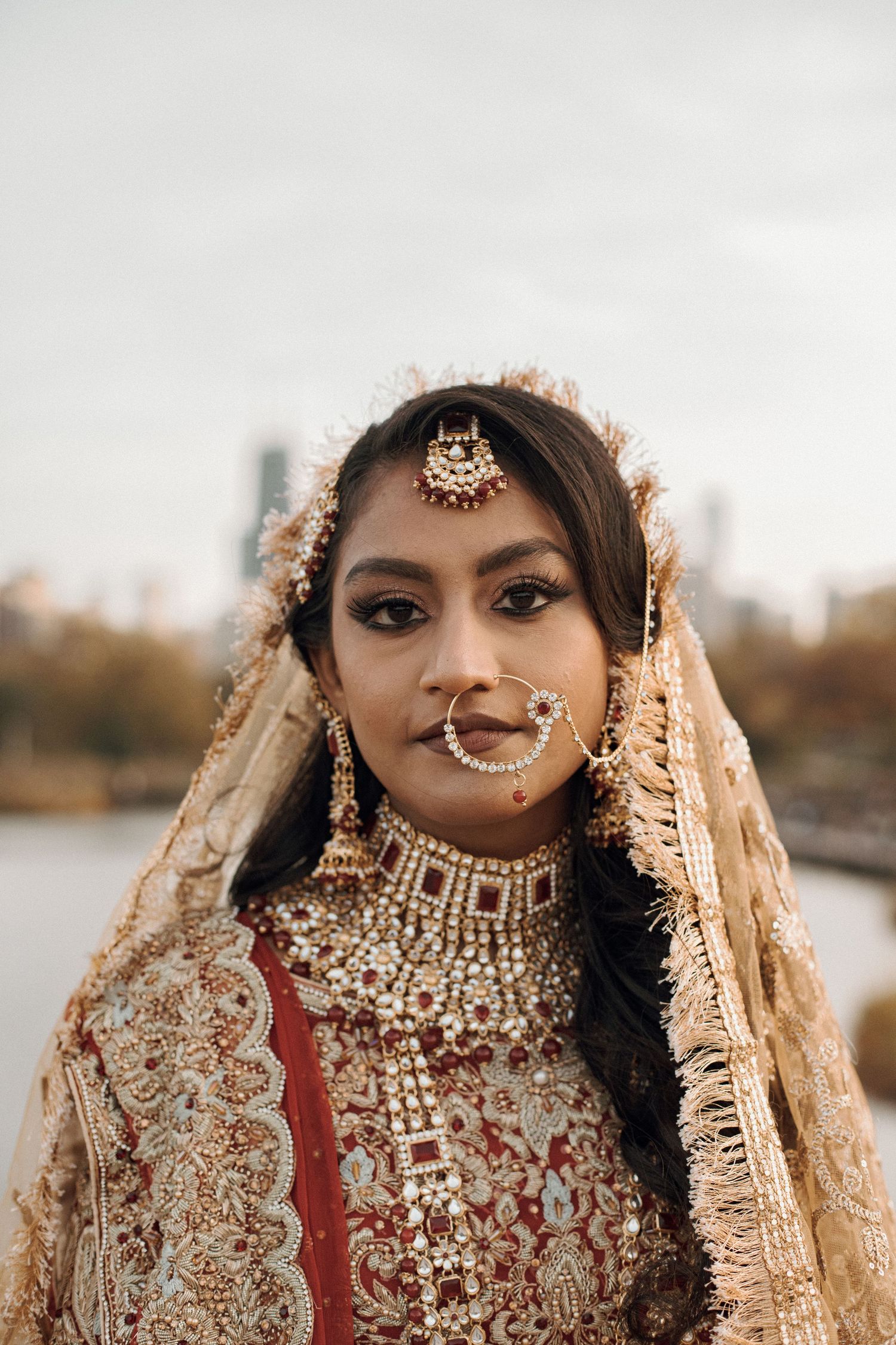 Portrait of a bride in elaborate gold jewelry, nose ring, maang tikka, and an embroidered red and gold veil outdoors.