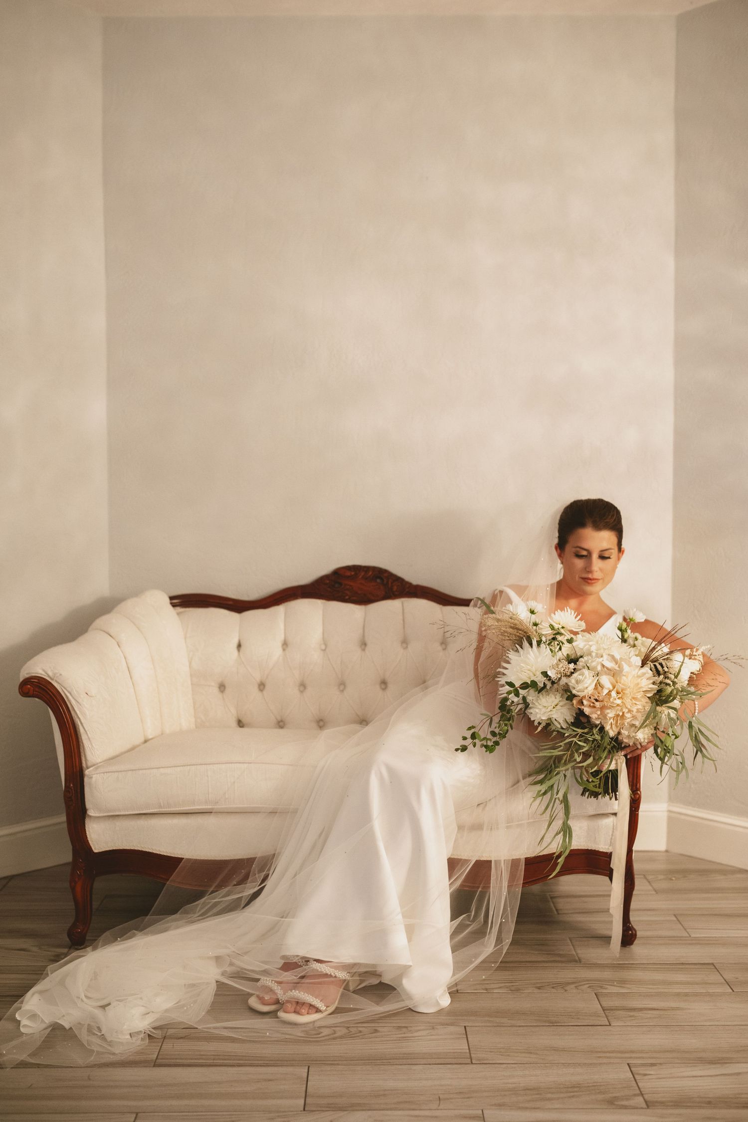 Bride in a white gown and long veil sits on a vintage tufted sofa holding a white and blush floral bouquet.