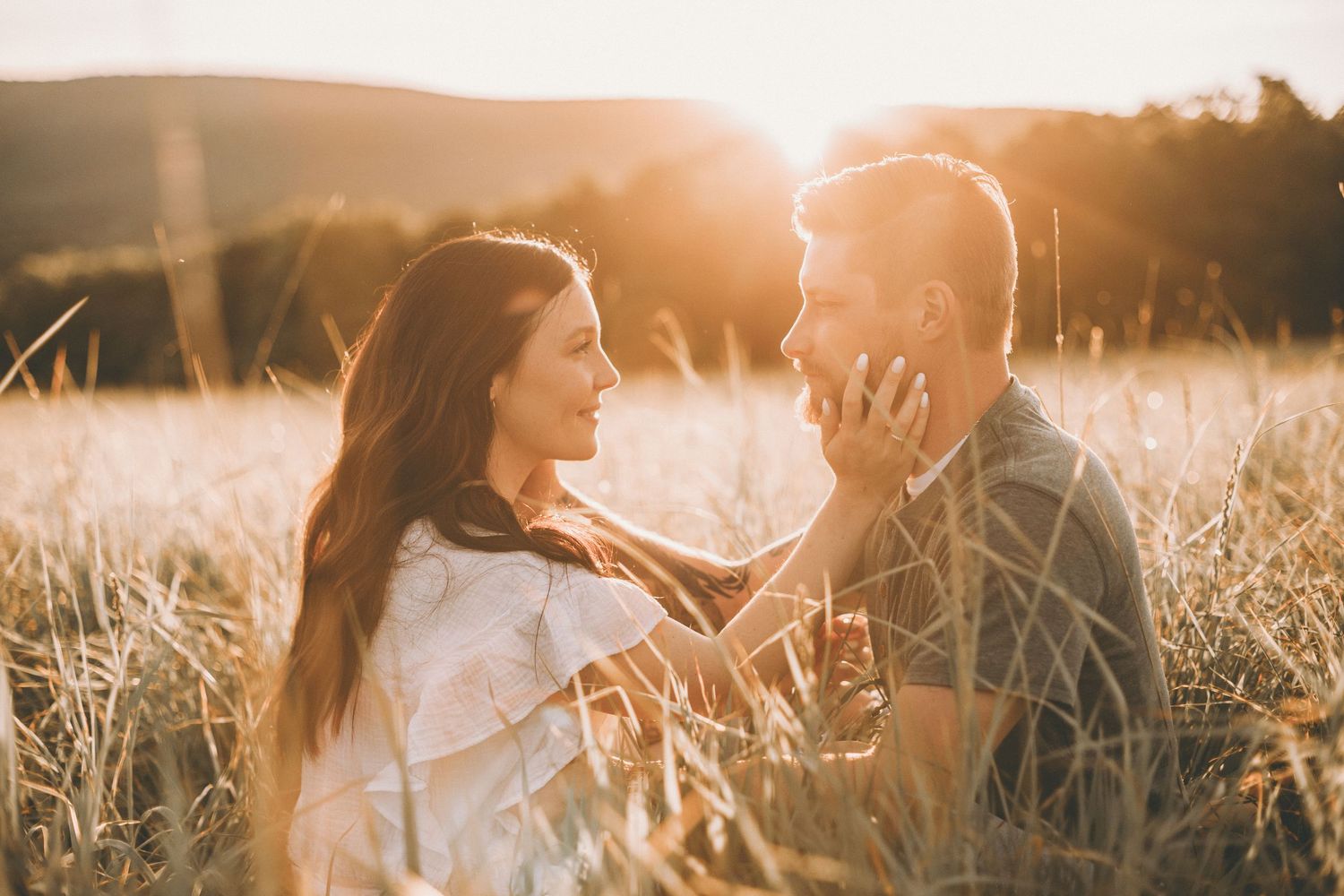 A smiling couple shares a tender moment in tall golden grass as warm sunset light glows behind them.