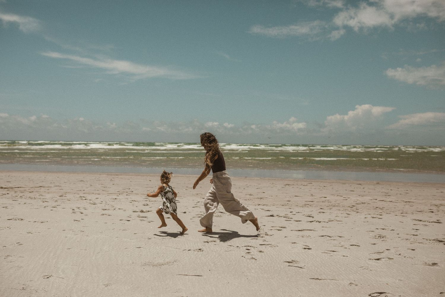 Mother and toddler running along a sandy beach toward the waves under a blue sky with scattered clouds.