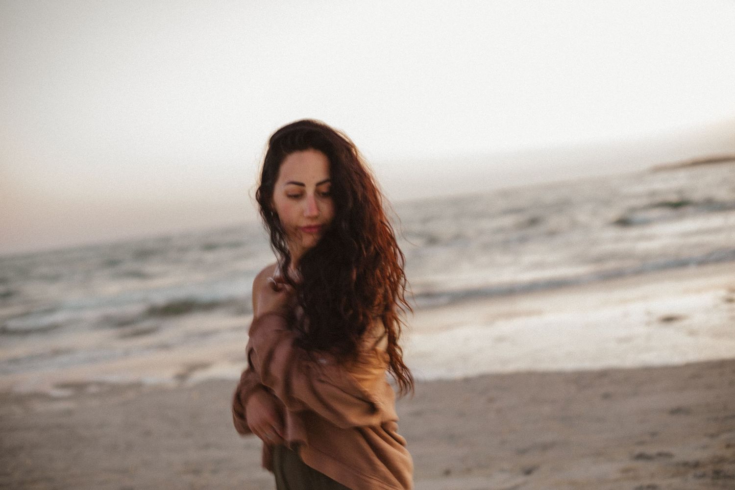Woman with long wavy dark hair wrapped in a brown shawl stands on a breezy beach at sunset.