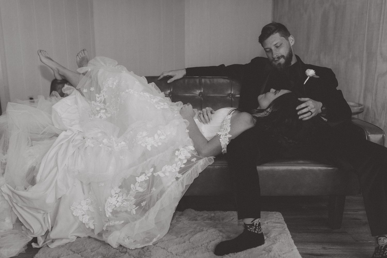 Black and white photo of a bride in a lace gown reclining across a leather couch with her groom in a dark suit.