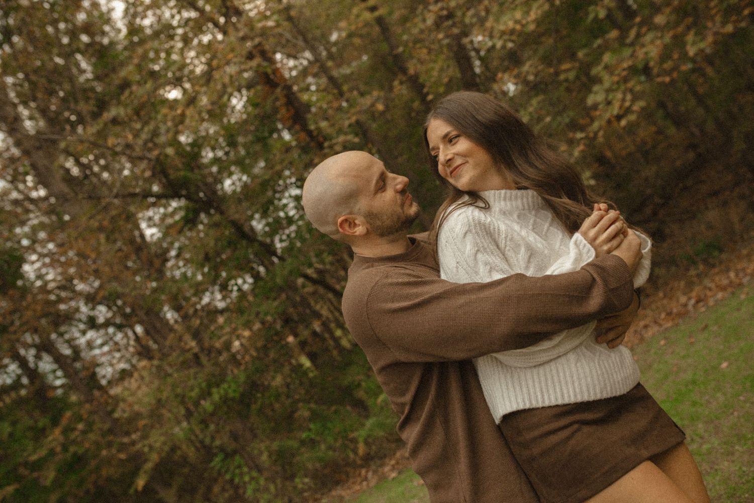 Bald man in a brown sweater embracing a smiling woman in a knit sweater among colorful autumn trees.