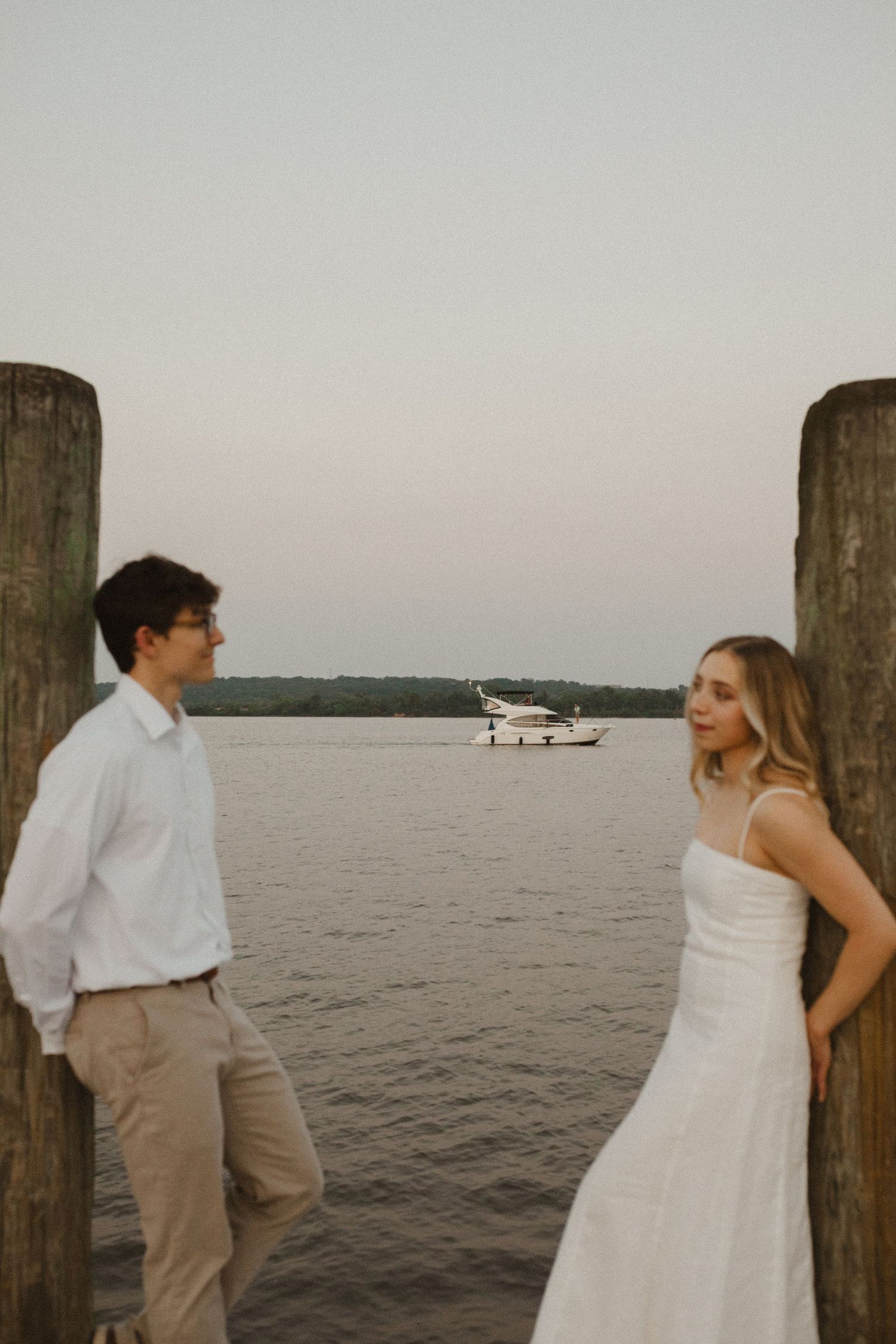 Couple leans against wooden dock posts facing each other with a boat floating on the calm river behind them.
