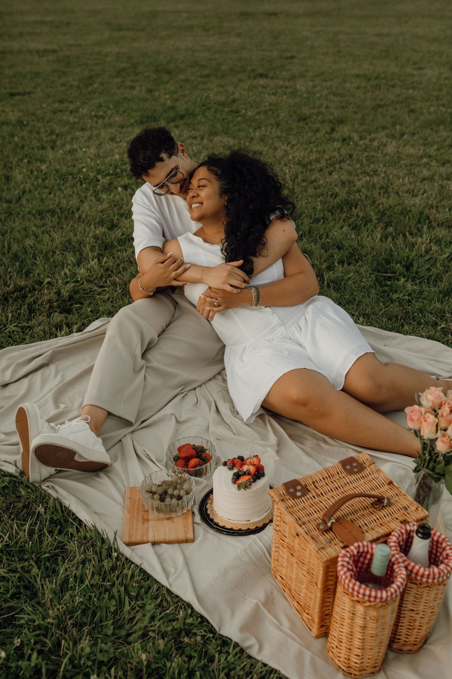A couple embraces and laughs on a picnic blanket in a grassy field with a wicker basket, cake and roses.