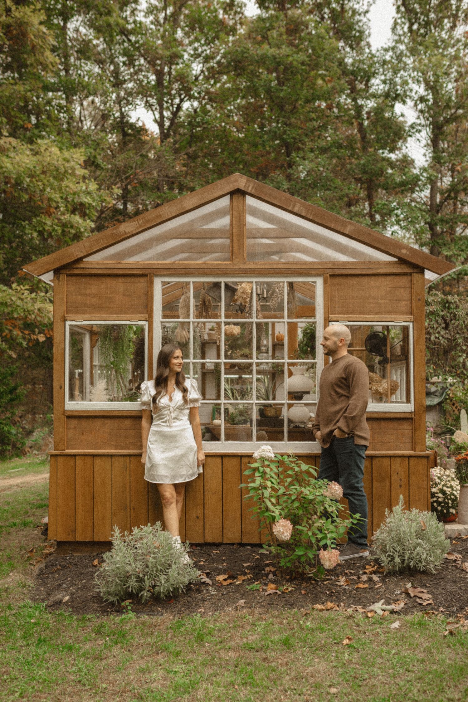 Couple stands beside a small wooden greenhouse framed by trees, flowers, and shrubs in a woodland engagement portrait.