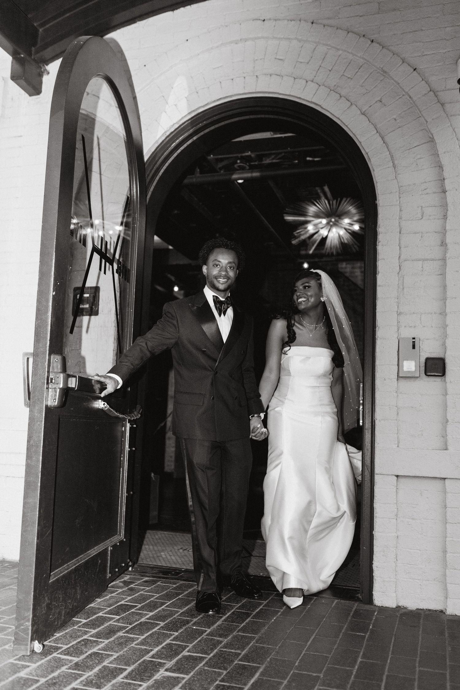 Black and white photo of a bride in a strapless gown and veil walking hand in hand with her groom through an arched brick doorway.