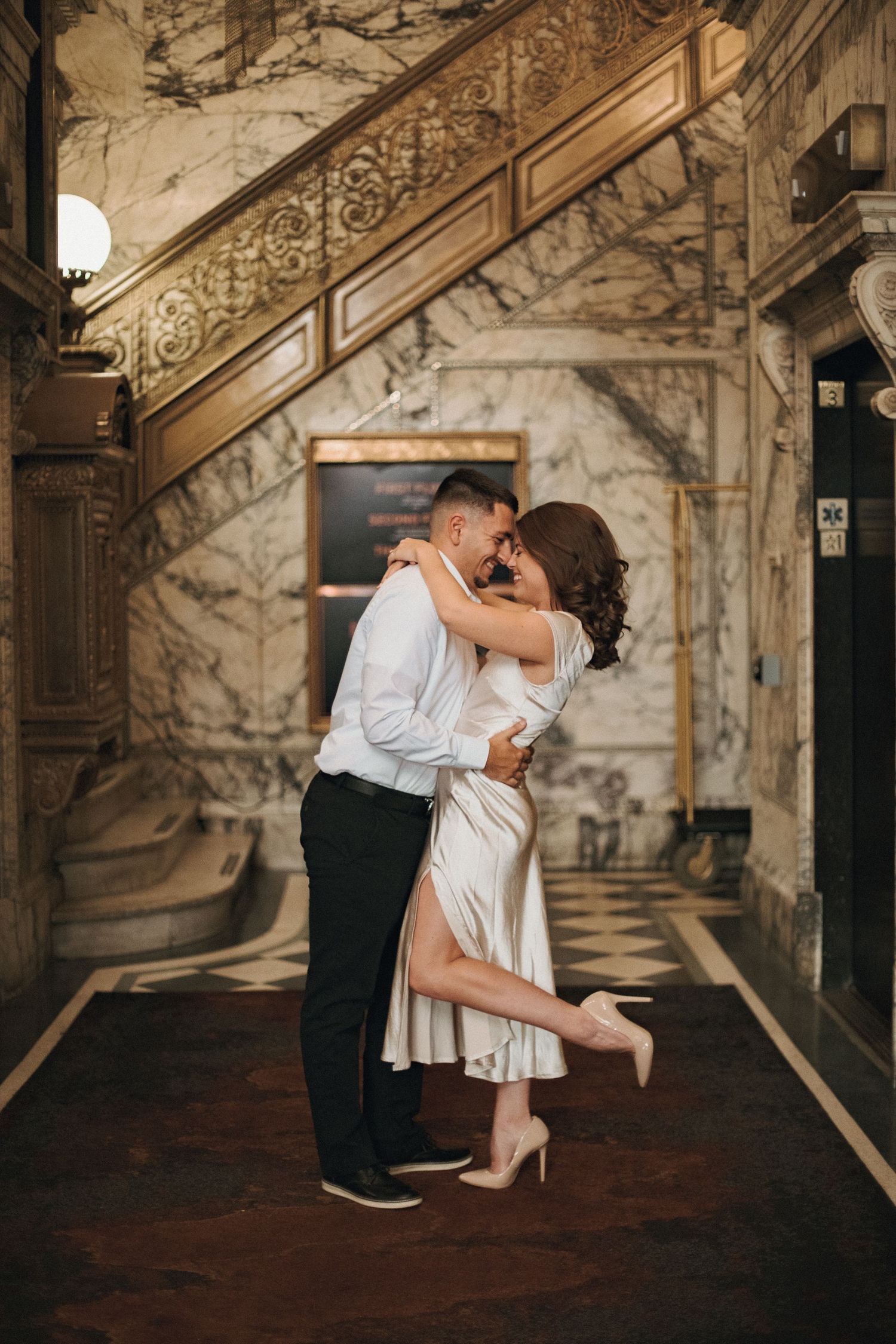 Couple embracing and kissing in an elegant marble lobby with an ornate staircase, the woman in a satin dress and heels.
