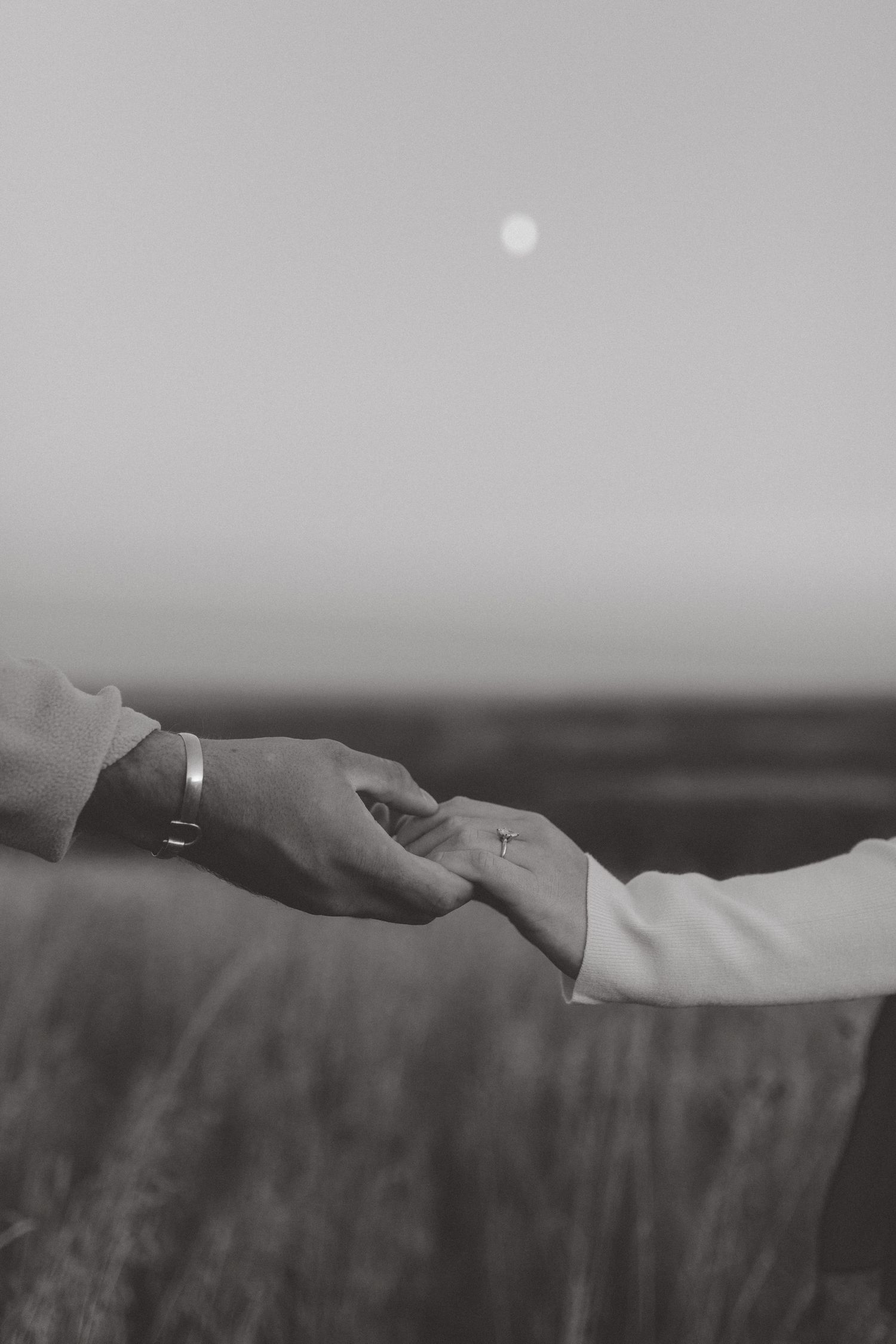 Black and white image of two hands gently held together in a field, with a bracelet visible and the moon in a hazy sky.