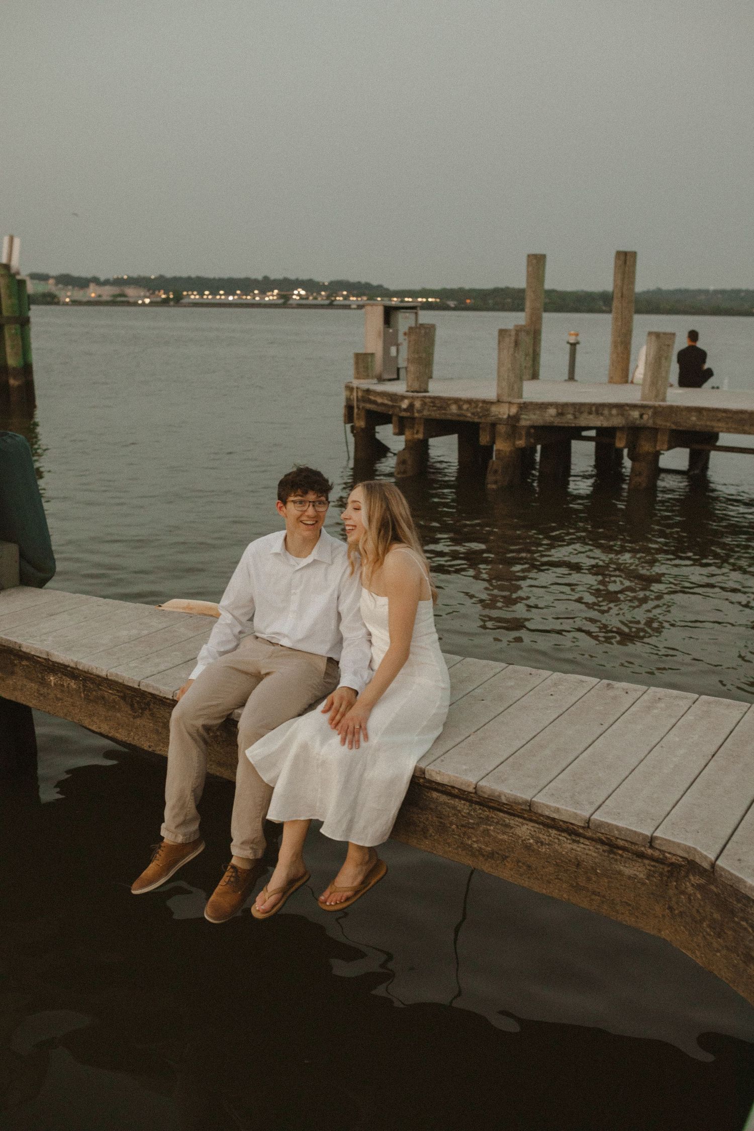 Smiling couple sits side by side on a wooden dock edge above the water at twilight during an engagement shoot.