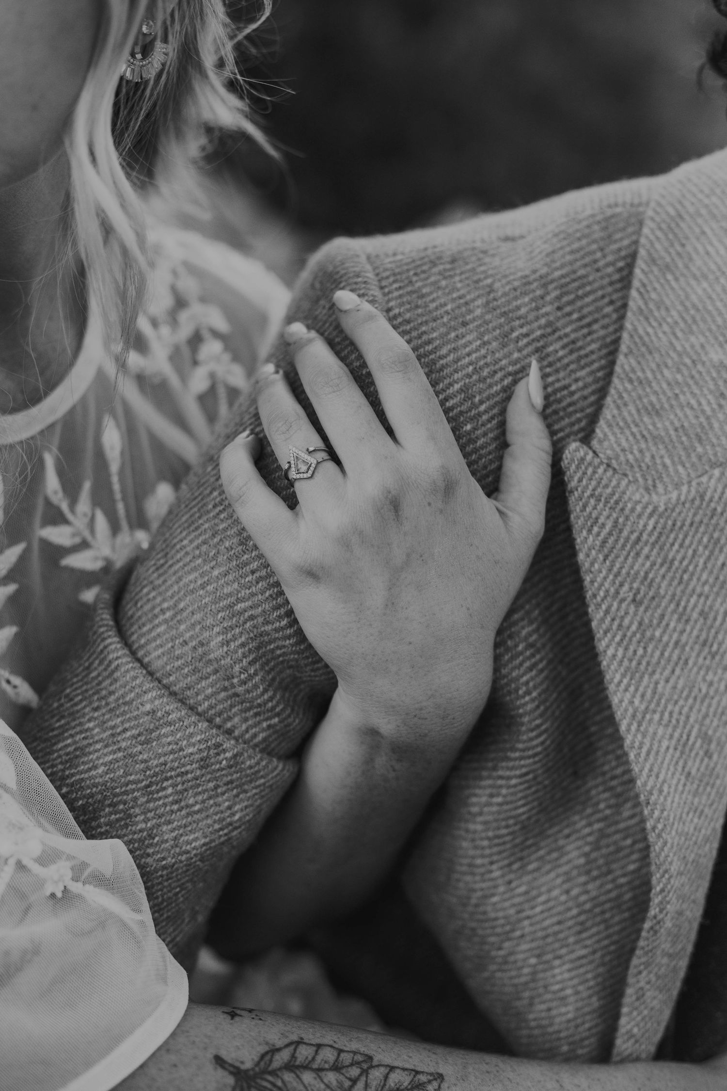 Black and white close-up of a bride's hand with a ring resting on her partner's shoulder in a wool jacket.