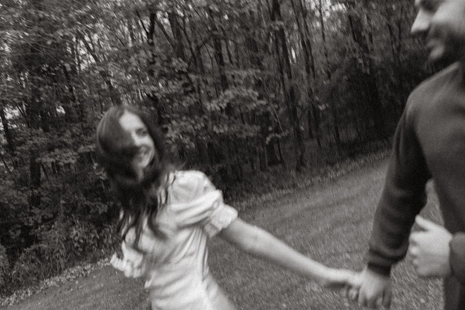 Black and white image of a laughing woman in a white dress holding a man's hand as they walk along a wooded path.