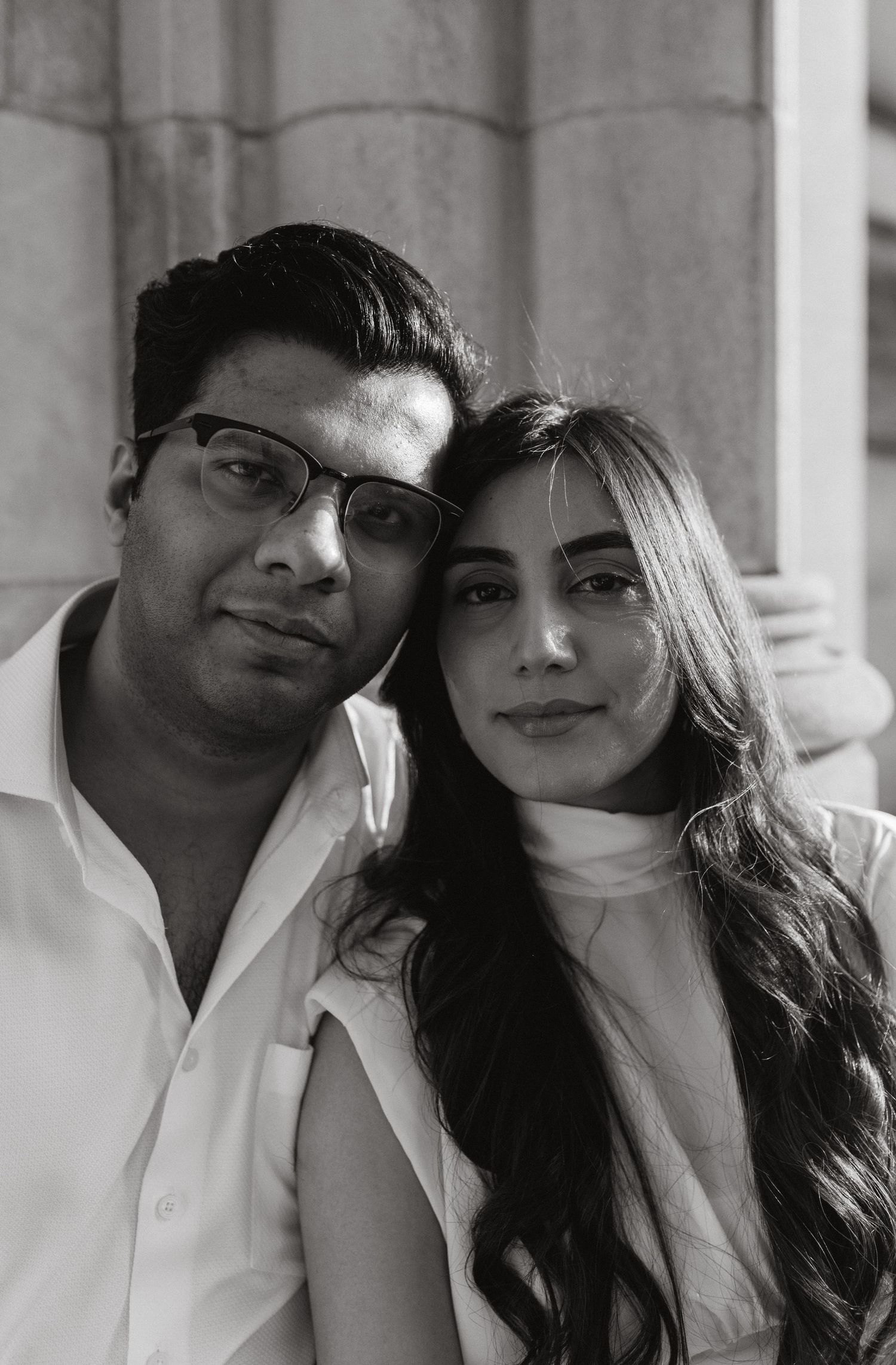 Black and white close-up portrait of a smiling couple leaning their heads together against a stone column.