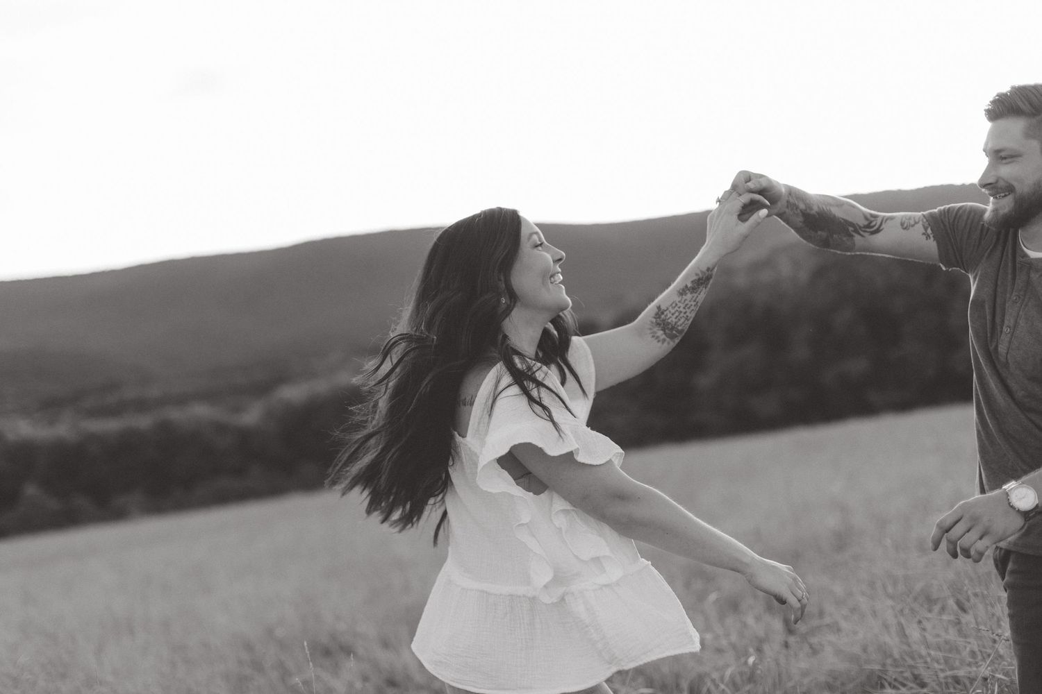 Black and white photo of a smiling couple dancing and twirling hand in hand in a grassy field with rolling hills behind them.