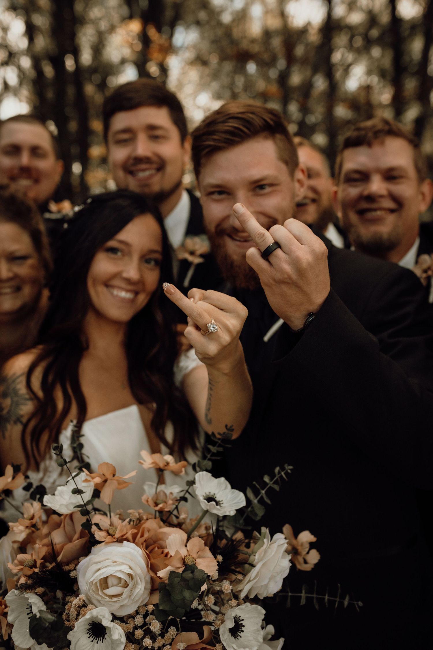 Smiling groom shows his wedding ring and points at his bride, with groomsmen behind them and a floral bouquet in front.