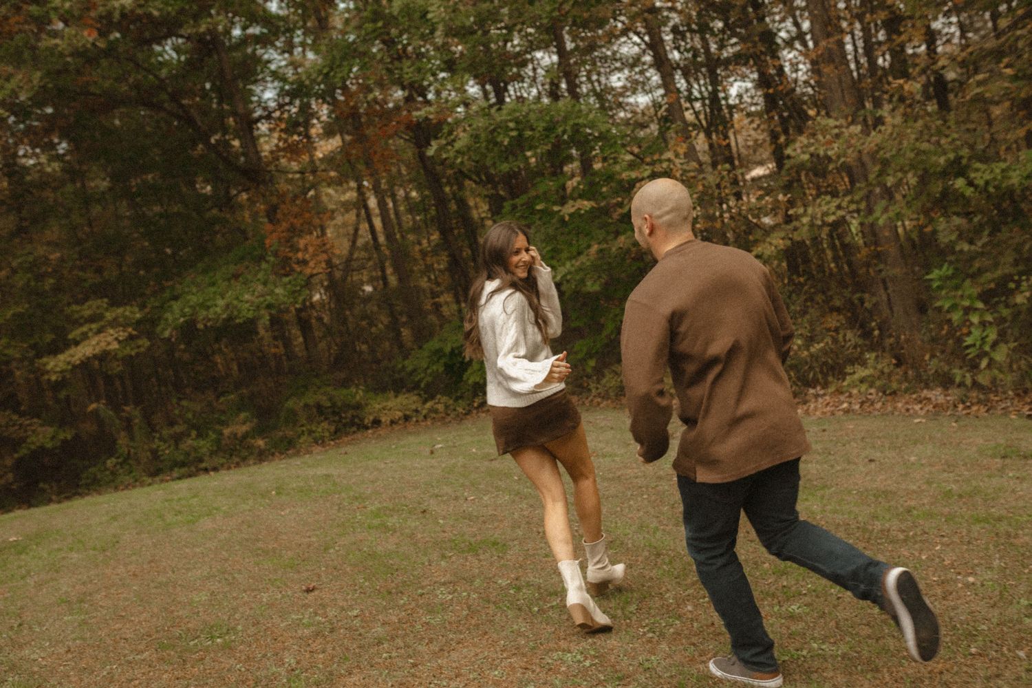 Couple running playfully across a grassy field beside a wooded area during an autumn photo shoot.