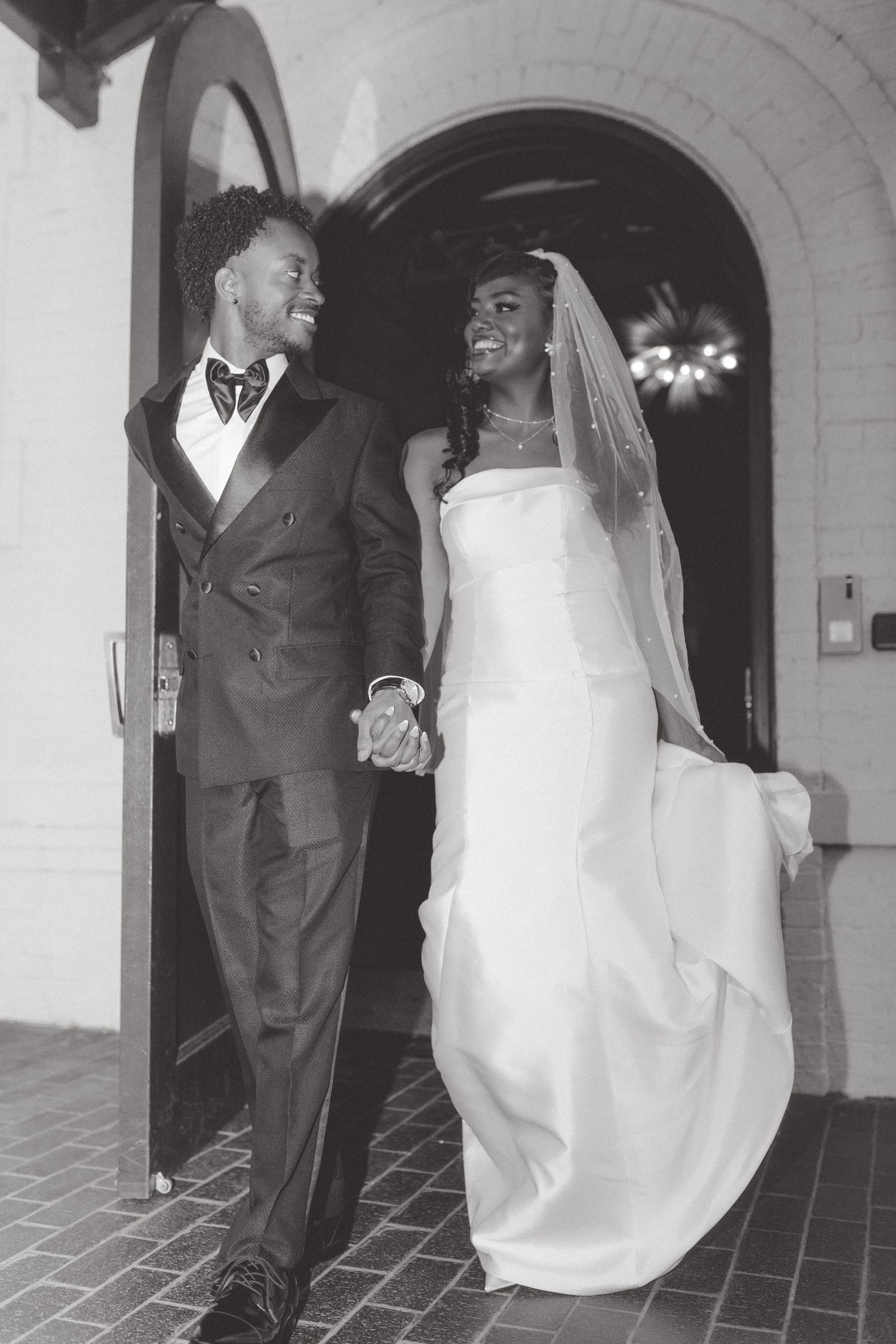 Black and white photo of a smiling bride and groom holding hands as they exit an arched doorway in a wedding gown and tuxedo.