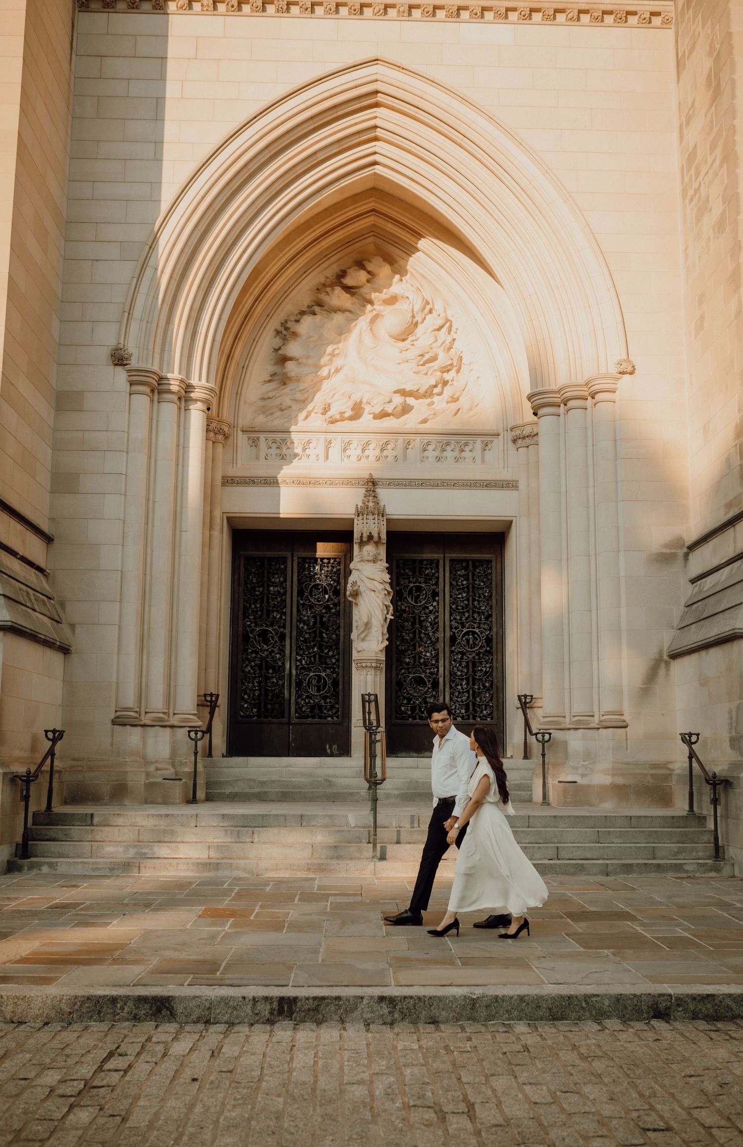 Couple walks hand in hand past the ornate arched entrance of a stone cathedral, the woman wearing a flowing white dress.