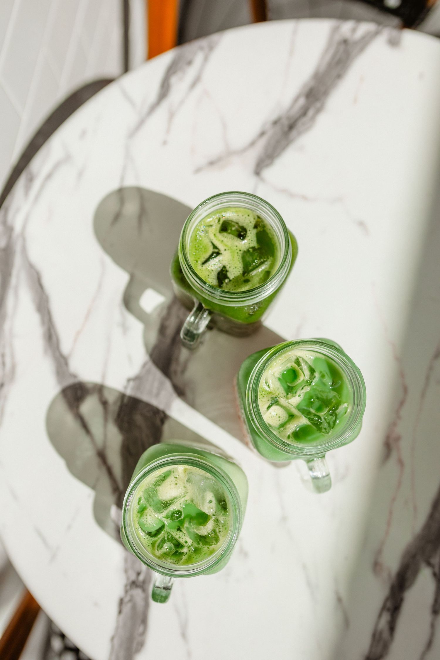 Overhead view of three iced matcha lattes in mason jar mugs on a marble table with sunlit shadows.