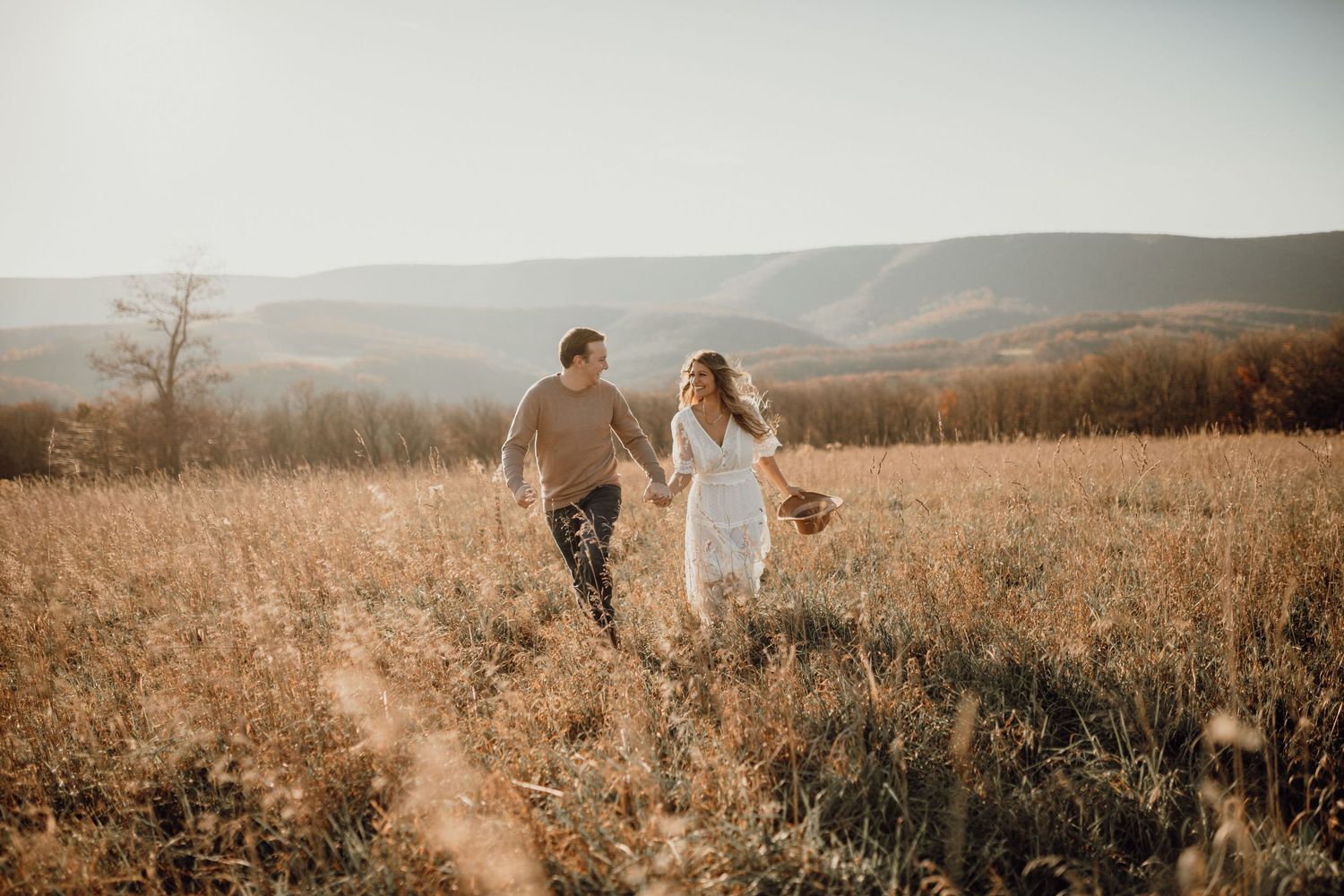 Couple holding hands and walking through a golden grassy field with rolling mountains in the background.