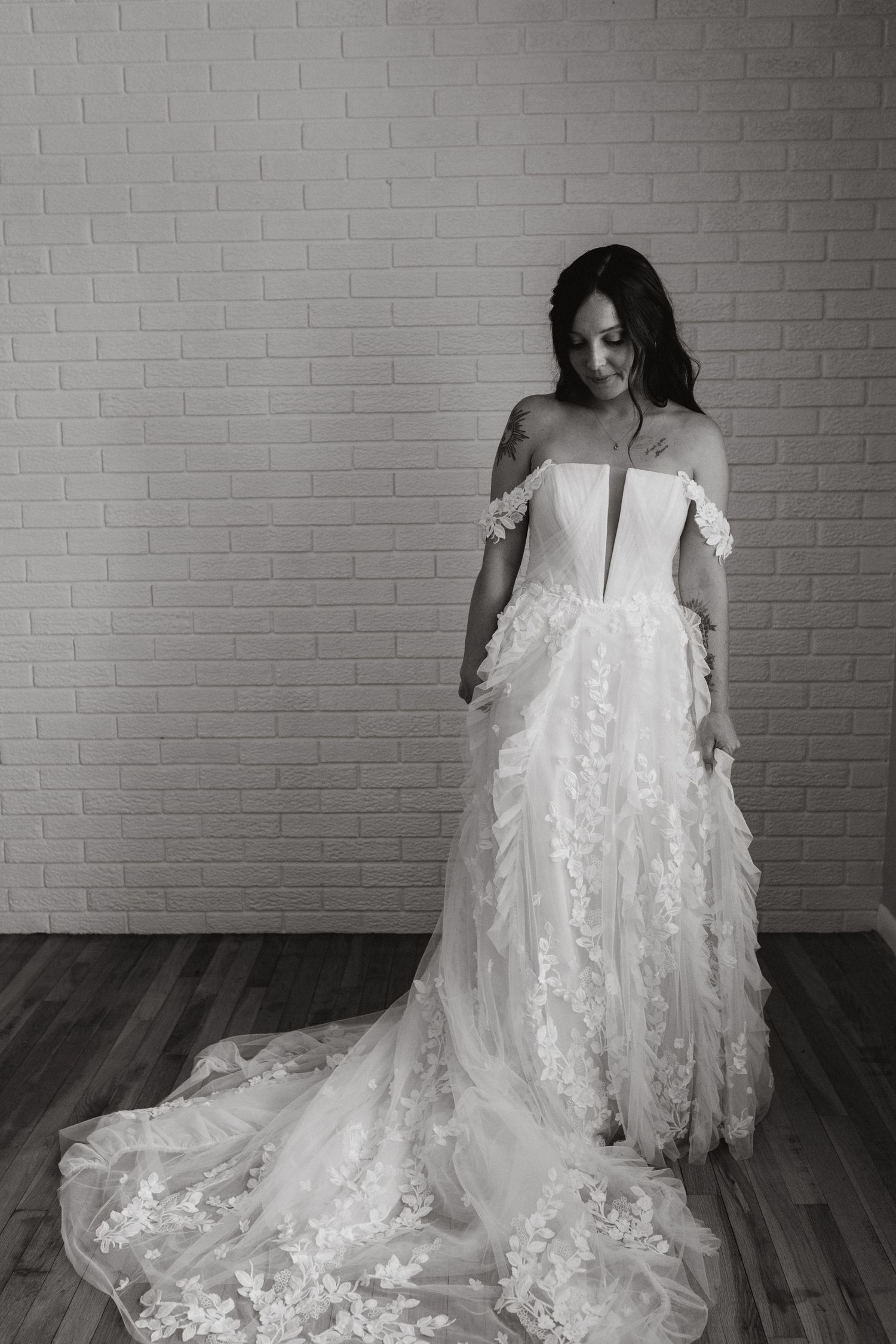 Black and white portrait of a bride in an off-shoulder floral lace wedding gown standing before a white brick wall.
