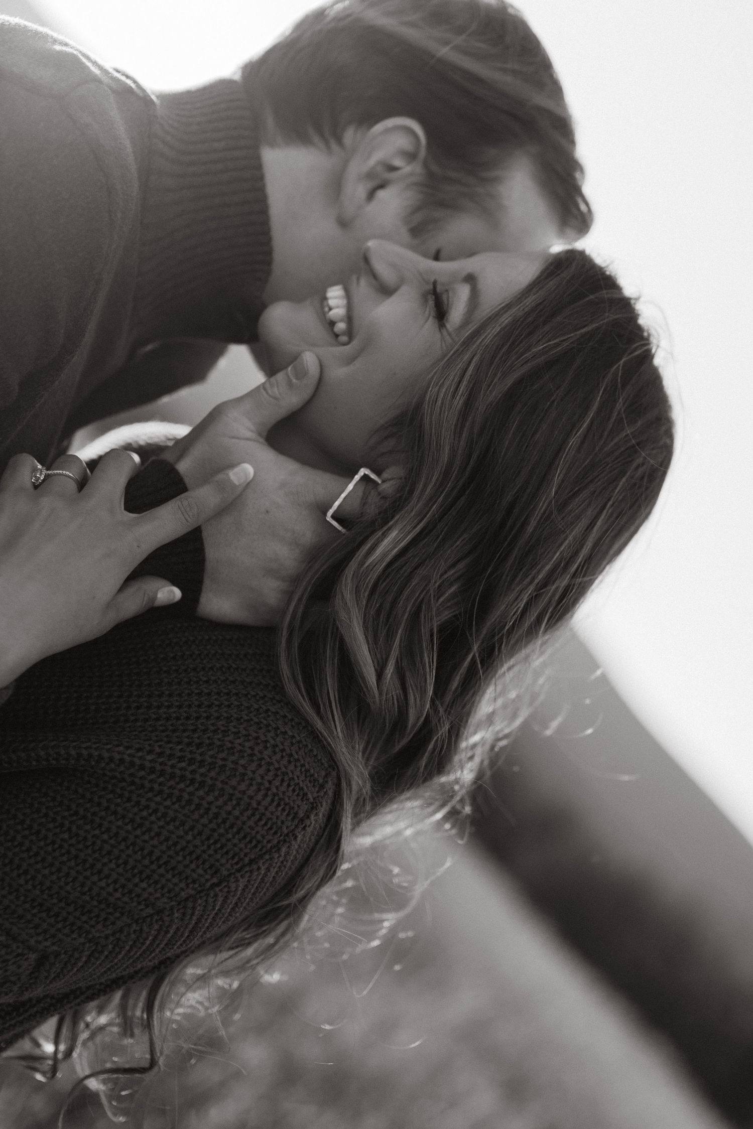 Black and white close-up of a man kissing the cheek of a laughing woman with long wavy hair.