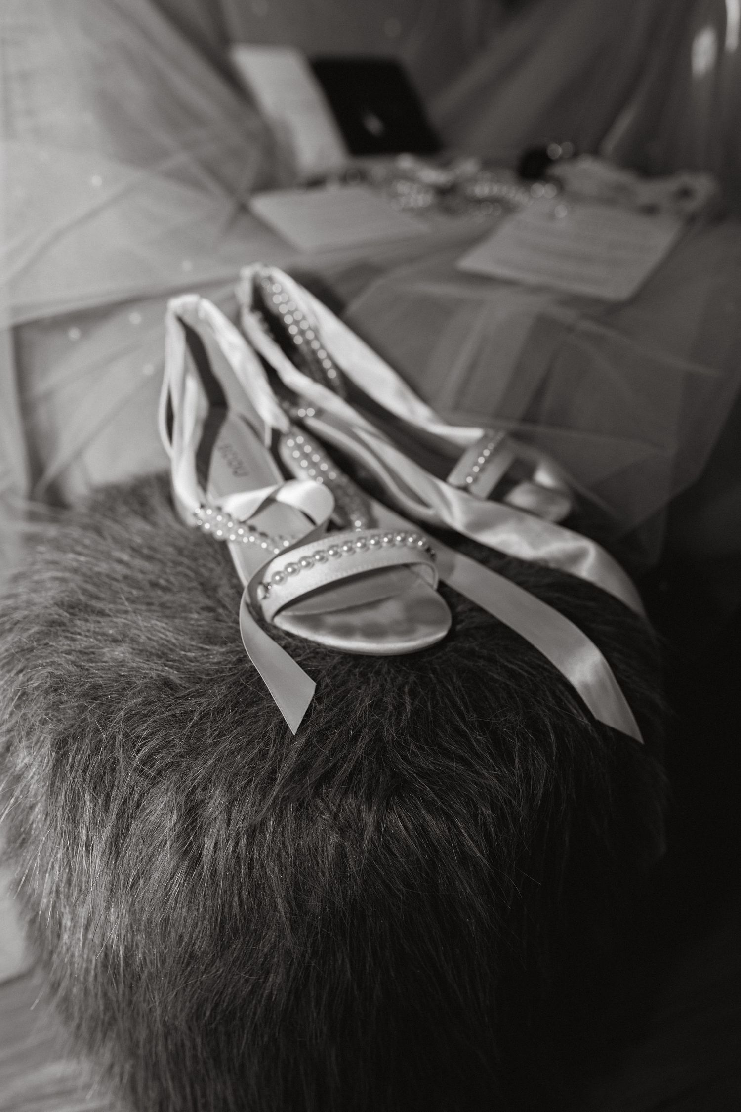 Black and white photo of beaded strappy bridal sandals with ribbon ties resting on a fur stool beside tulle.