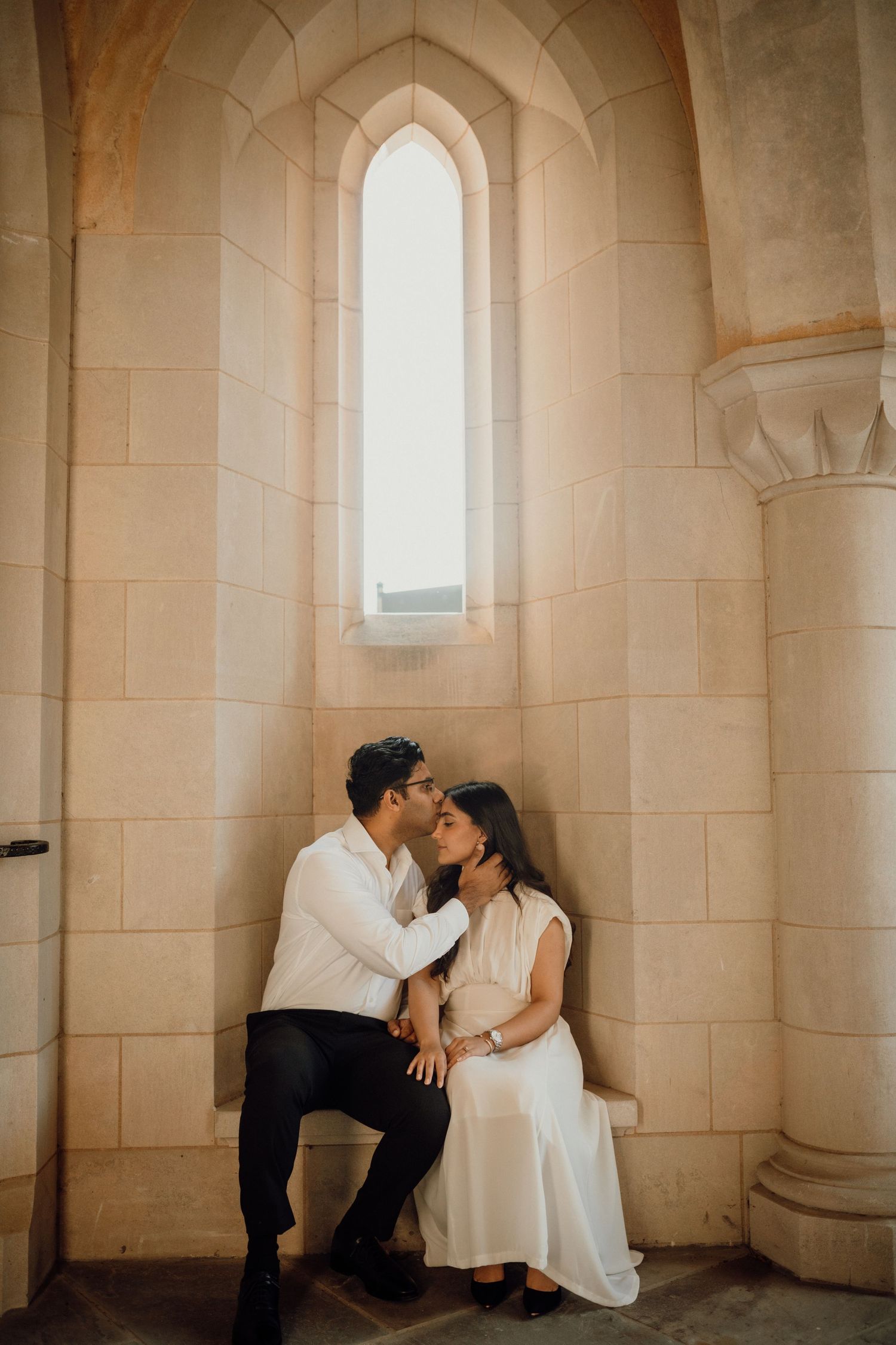 Expecting couple sits together in a stone alcove beneath a tall arched window, touching foreheads in a tender maternity moment.