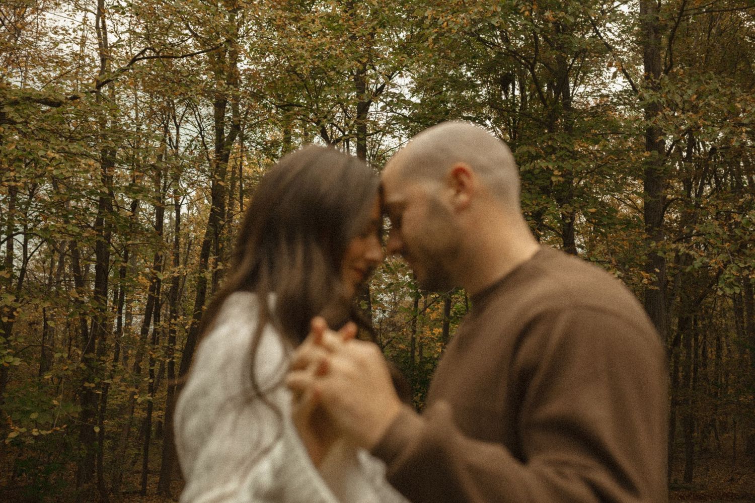 Couple standing forehead to forehead and holding hands in an autumn forest during an engagement photo session.