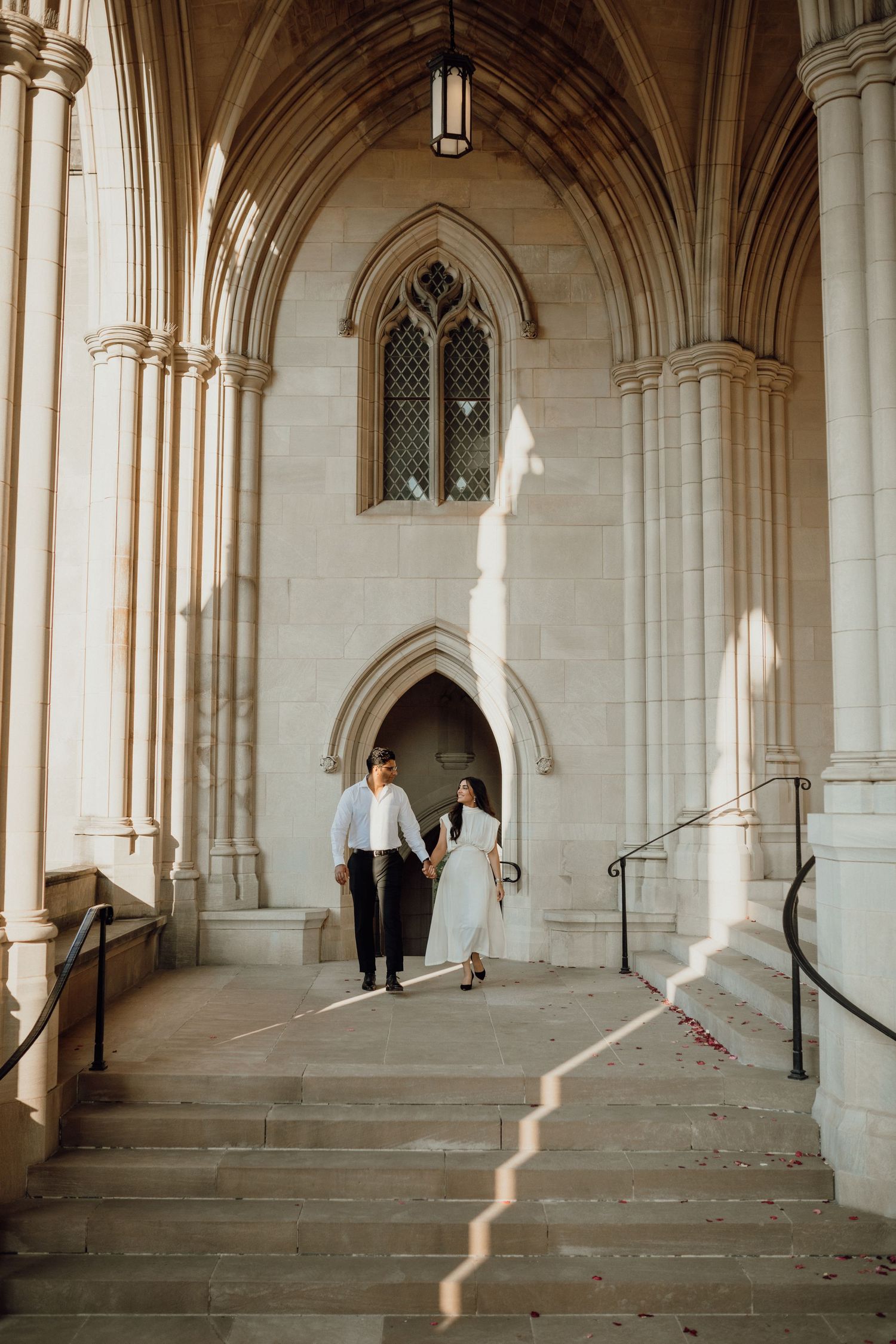 Couple in white and black walk hand in hand along a sunlit Gothic stone corridor during an engagement photo session.