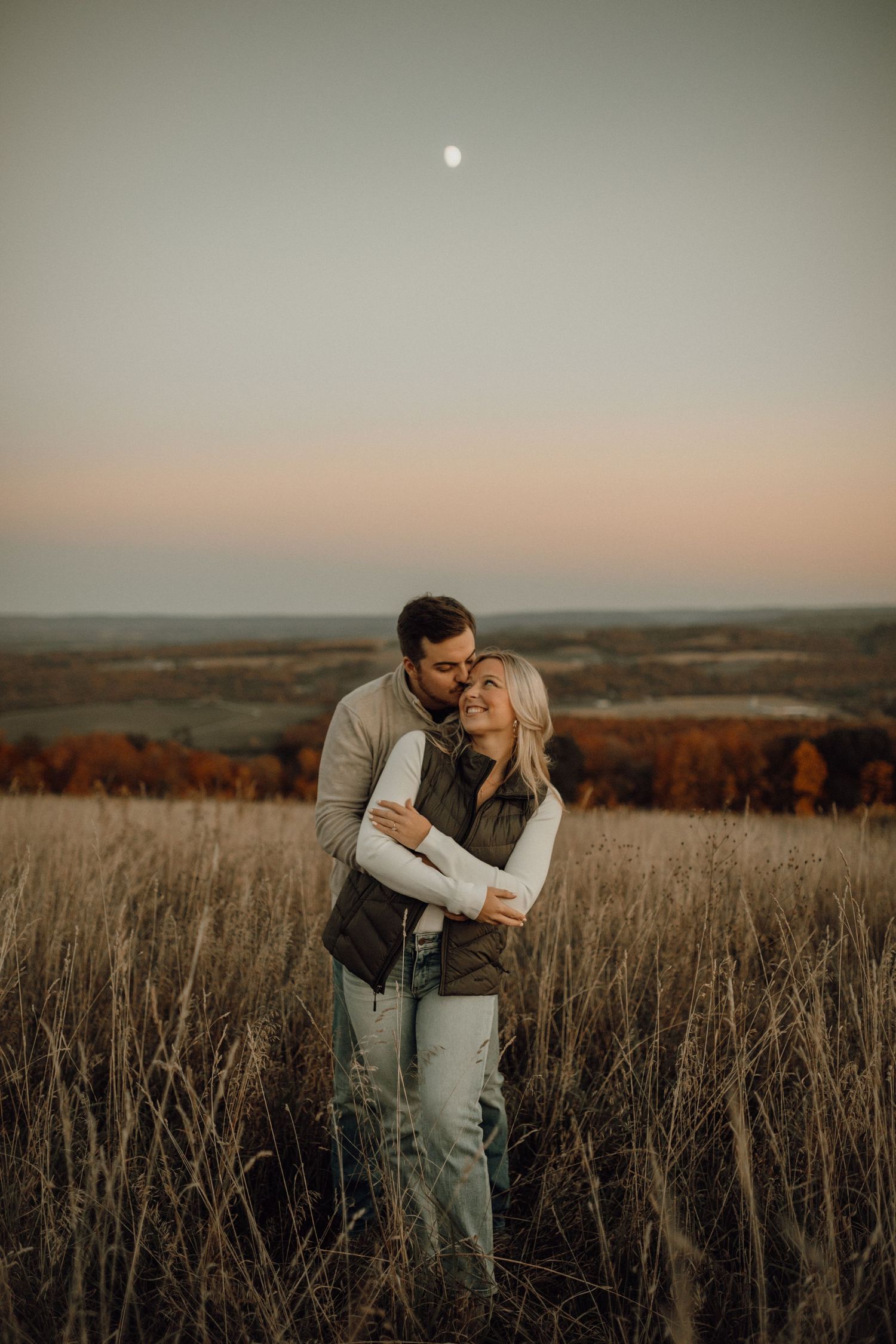 A smiling man kisses a woman's forehead as he hugs her from behind in a grassy field with autumn trees and a moonlit dusk sky.