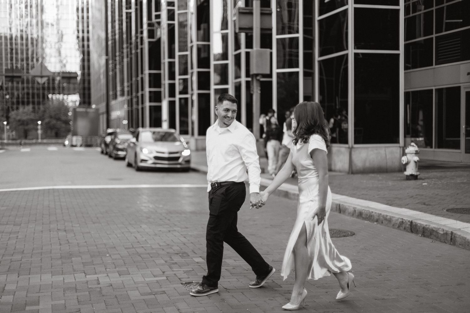 Couple holding hands walking across a city street past glass skyscrapers in a black and white engagement photo.