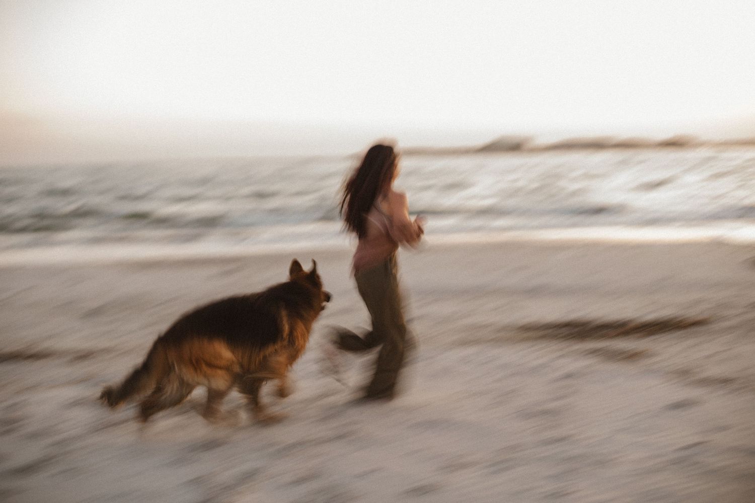 Blurred motion photo of a woman running on a beach at sunset with a German Shepherd beside her near the waves.