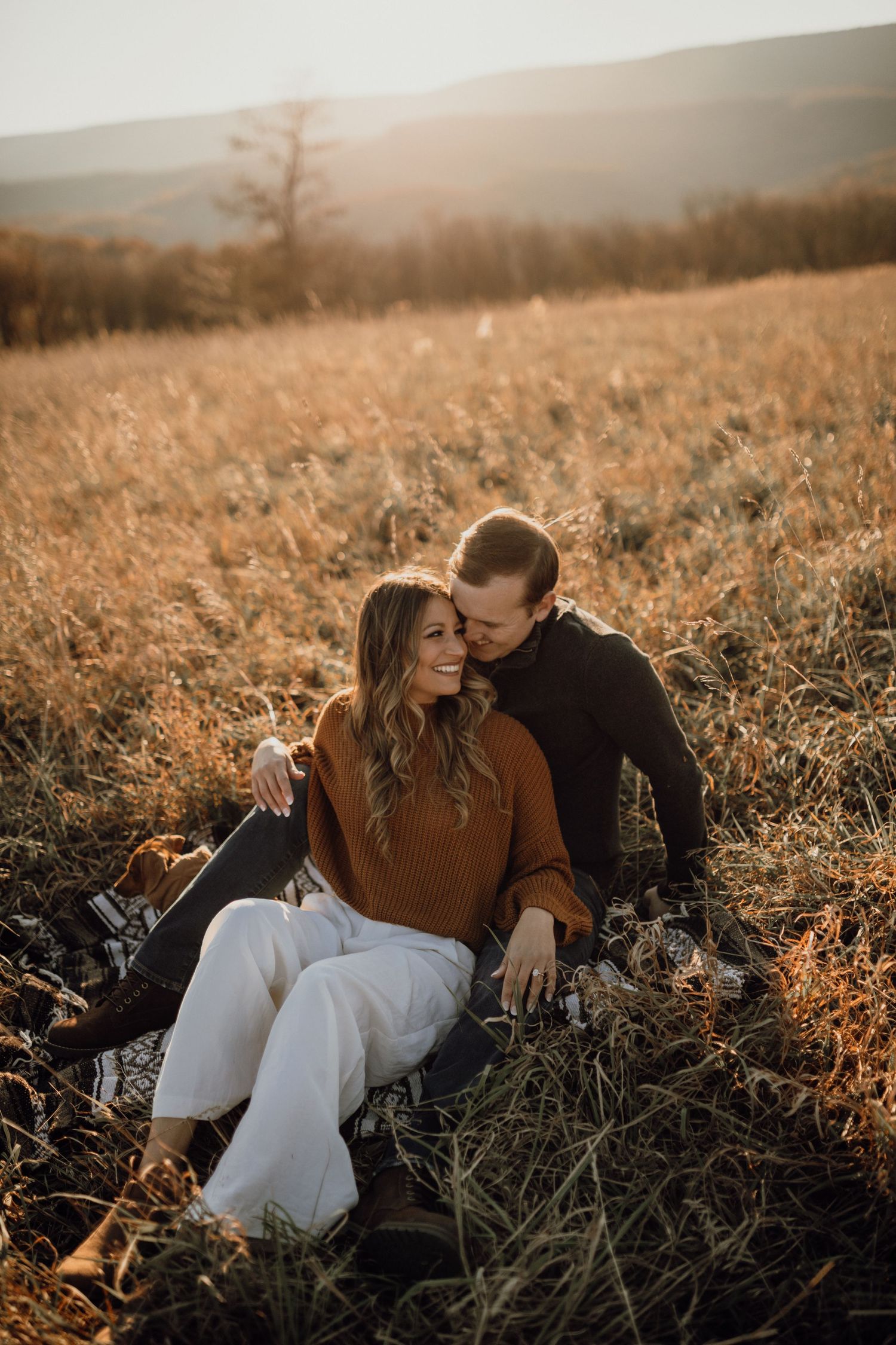 Smiling couple cuddling on a blanket in tall golden grass during an engagement photo session at sunset.