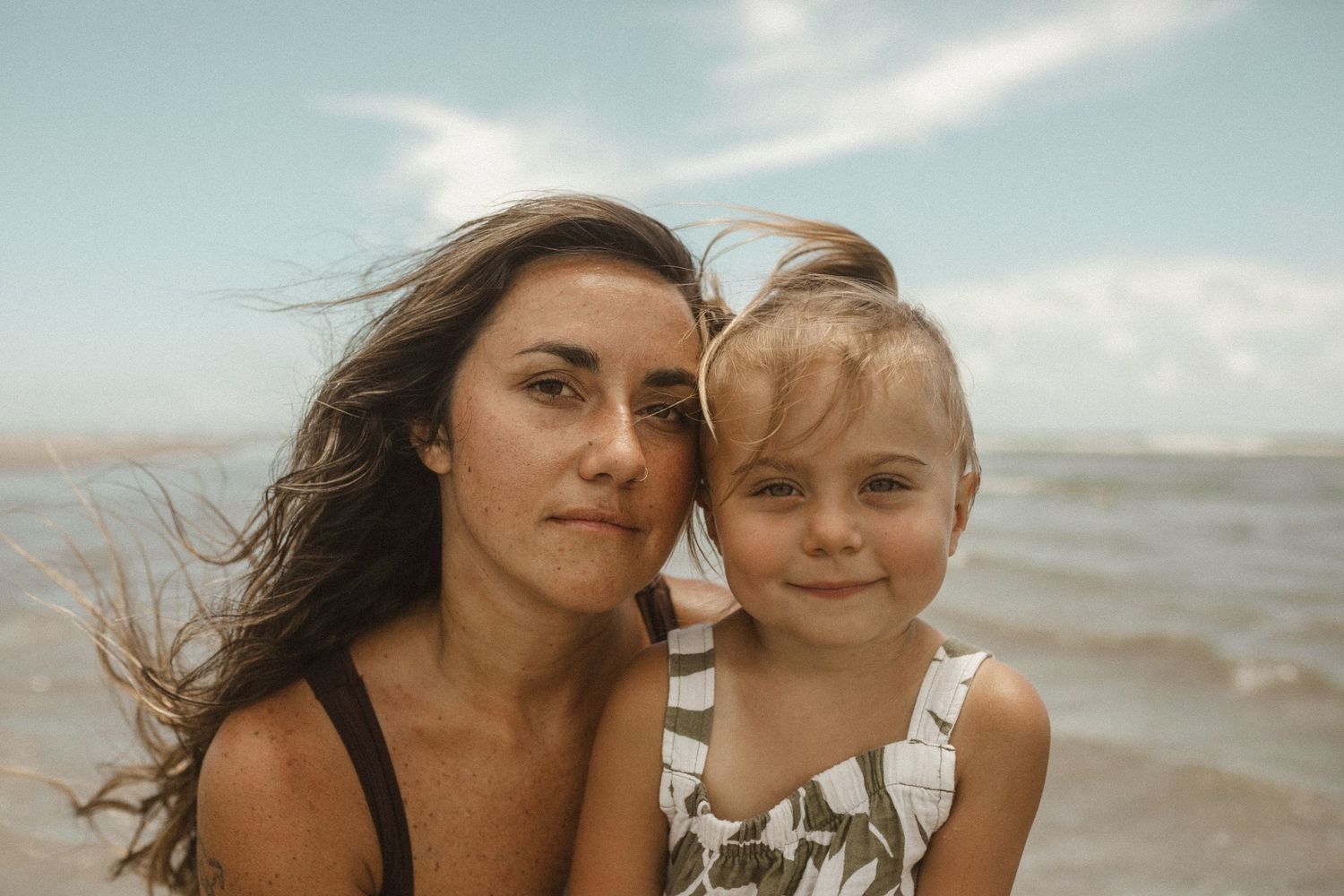 Mother and young daughter pose cheek to cheek on a breezy beach with the ocean and cloudy sky behind them.