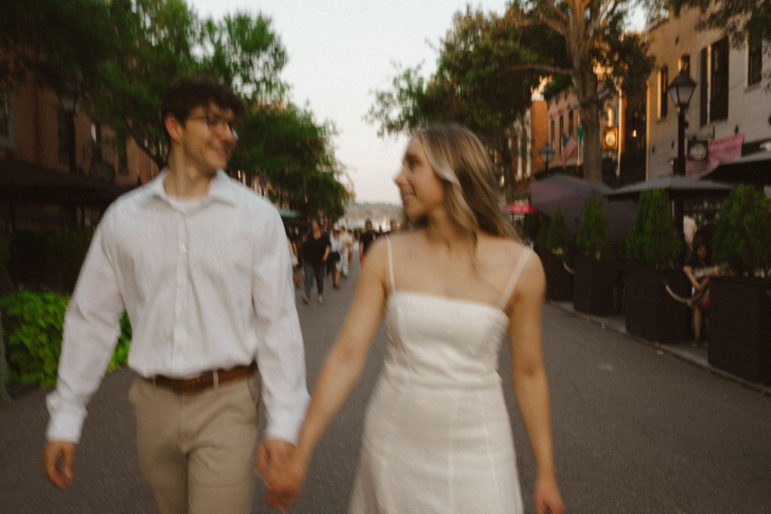 Couple in white and khaki holds hands and walks down a charming city street at golden hour for engagement photos.