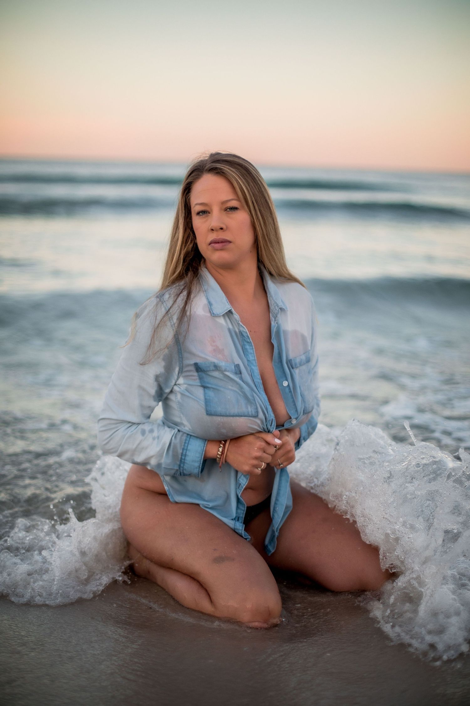 Blonde woman in open denim shirt kneeling in ocean waves at sunset on sandy beach. Cape cod beach Boudoir photos