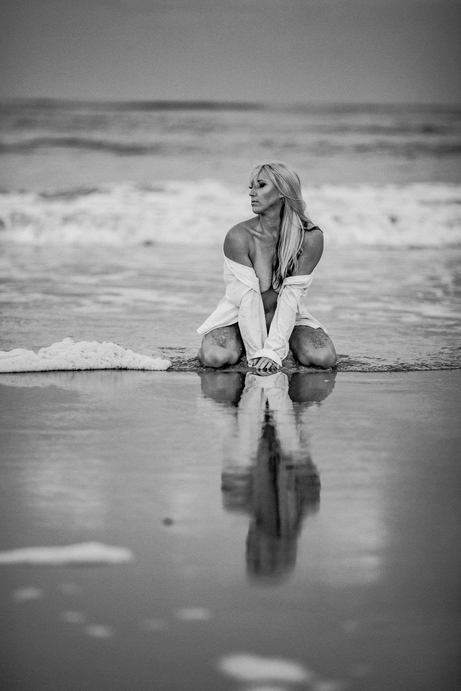 Black and white artistic photo of blonde woman kneeling at ocean shoreline with reflection. Boudoir photos