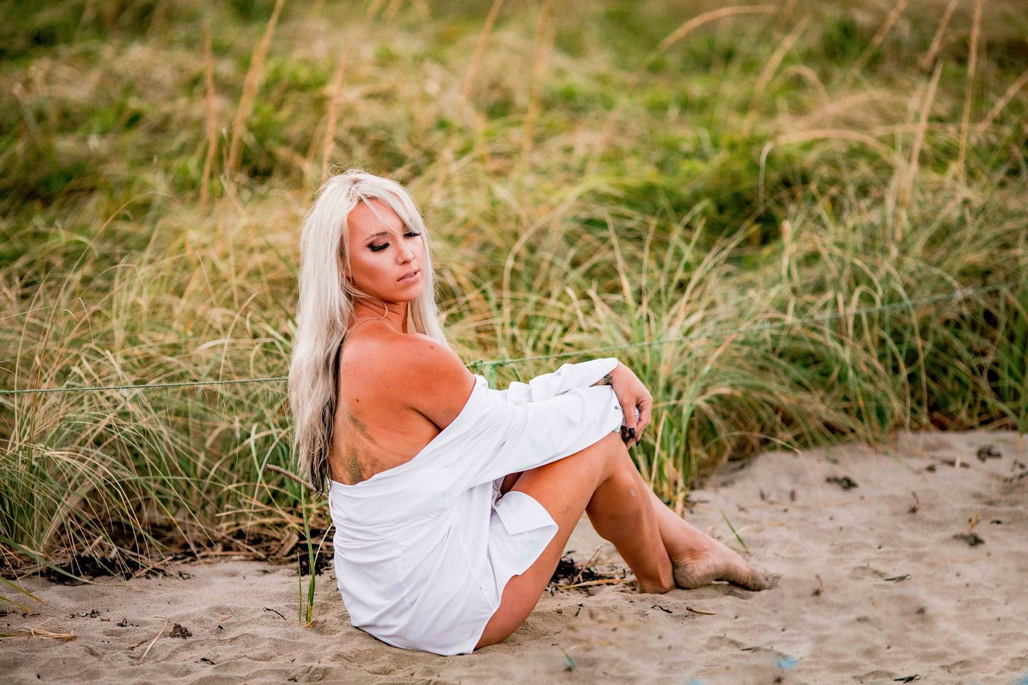 Blonde woman in white wrap sitting on sandy dunes surrounded by tall grass Cape cod beach Boudoir photos
