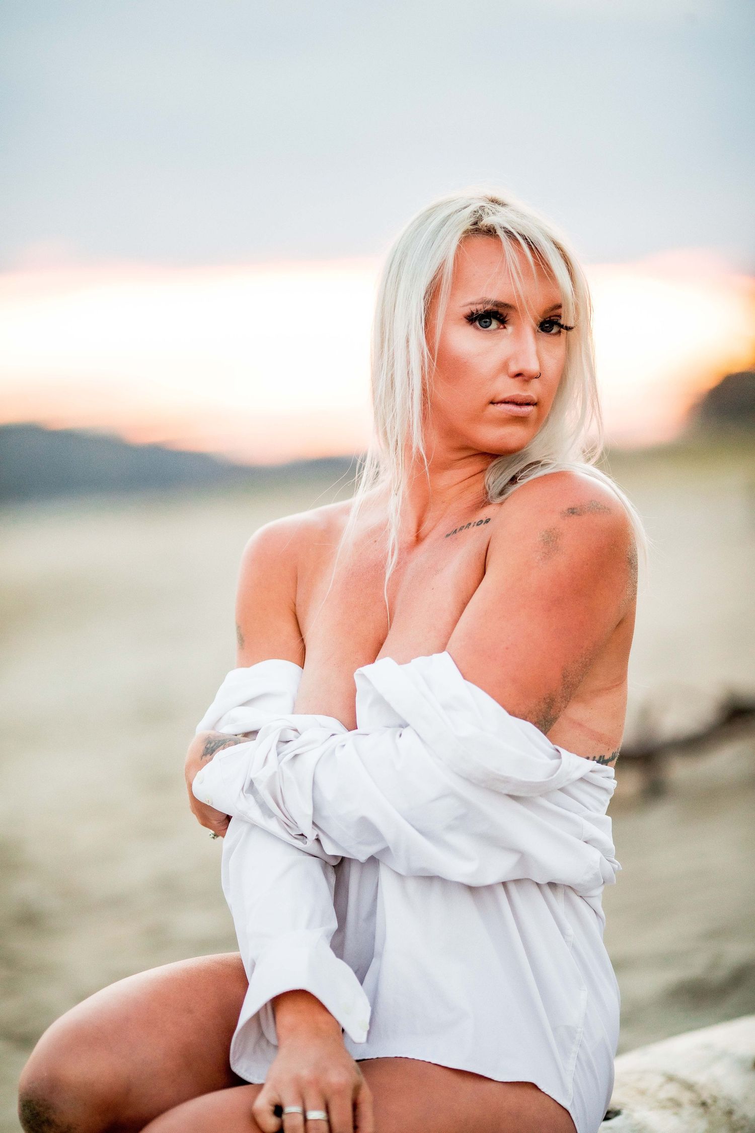 Blonde woman in white off-shoulder shirt posing on desert sand during colorful sunset. Cape cod beach Boudoir photos