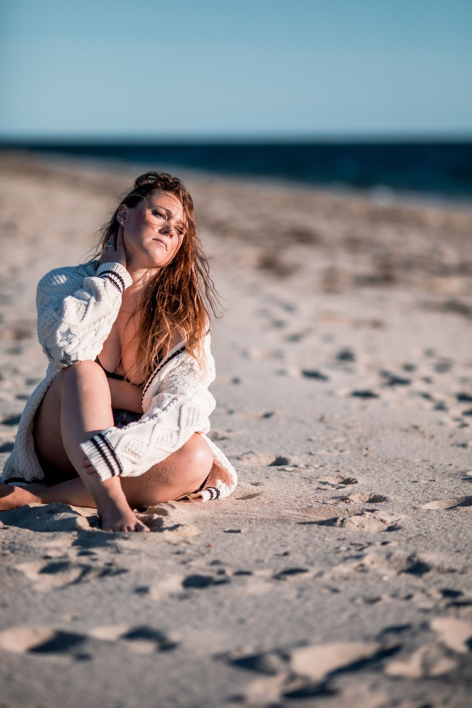 Woman in white knit cardigan and bikini sitting on sandy beach in sunlight. Cape cod beach Boudoir photos