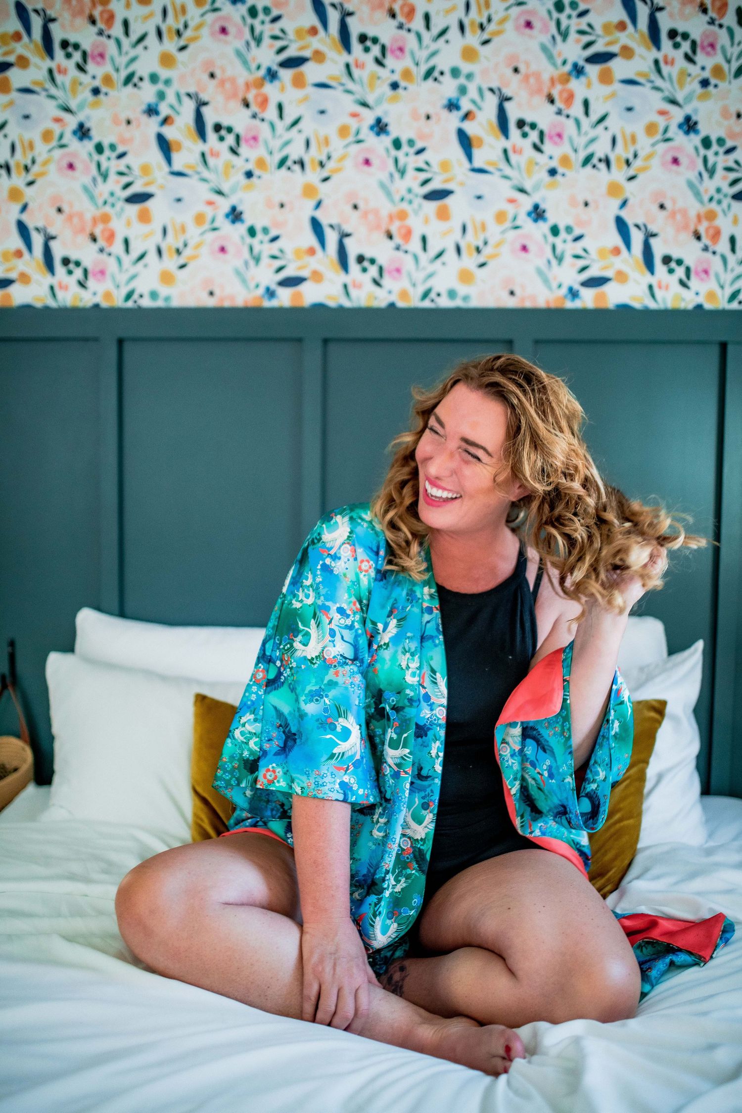 Smiling woman with curly hair sitting on bed wearing teal floral robe with colorful wallpaper background Cape cod studio Boudoir photo