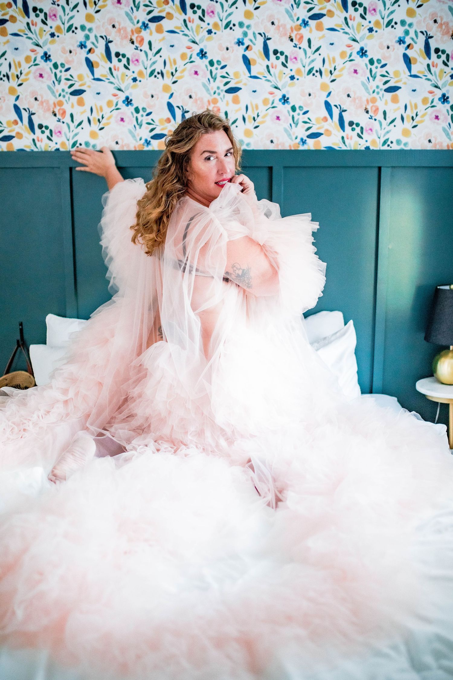 Woman in voluminous pink tulle robe posing playfully on white bed with floral wallpaper backdrop. Cape cod beach Boudoir photos