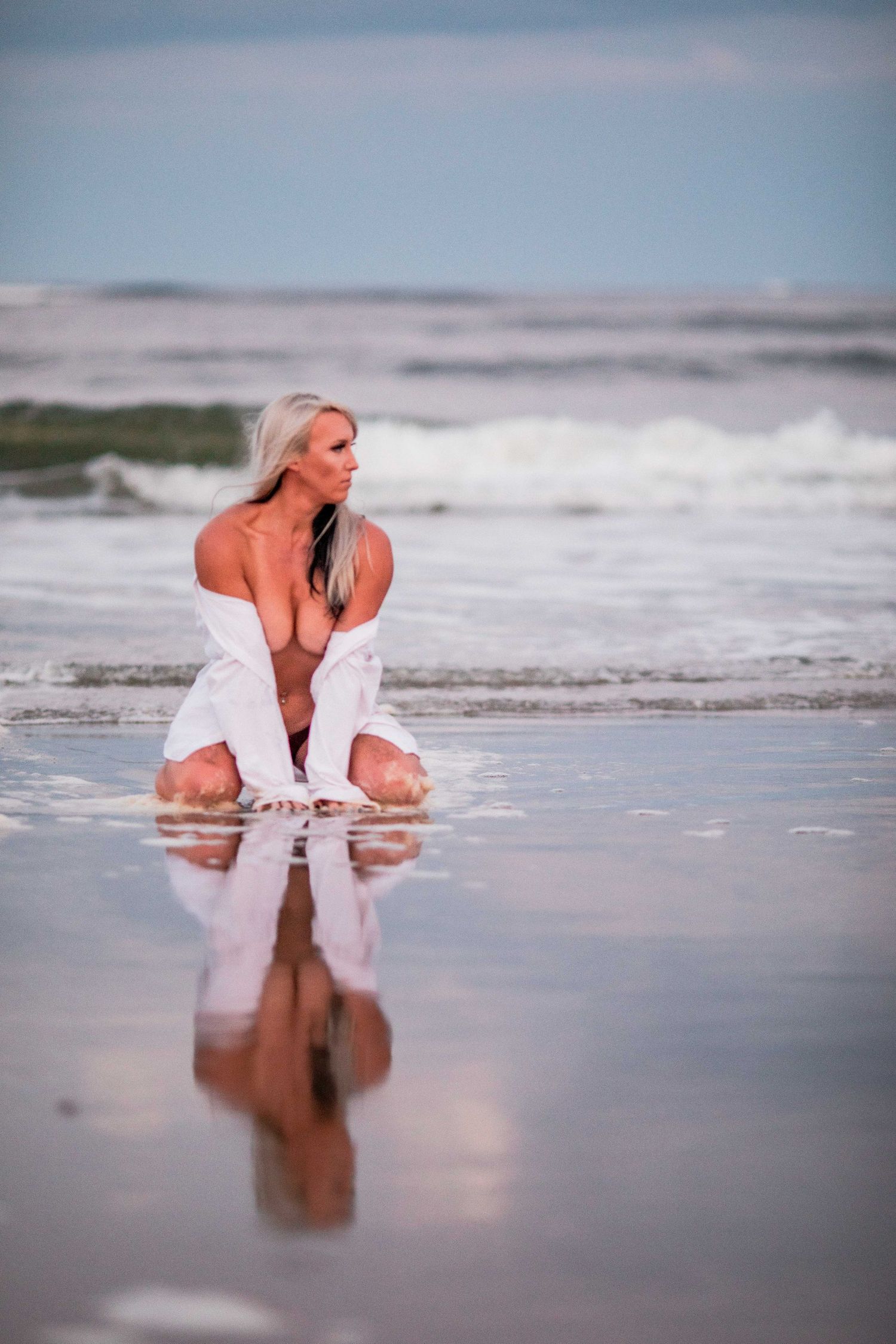 Blonde woman in open white shirt kneeling on reflective wet beach near ocean waves. Cape cod beach Boudoir photos