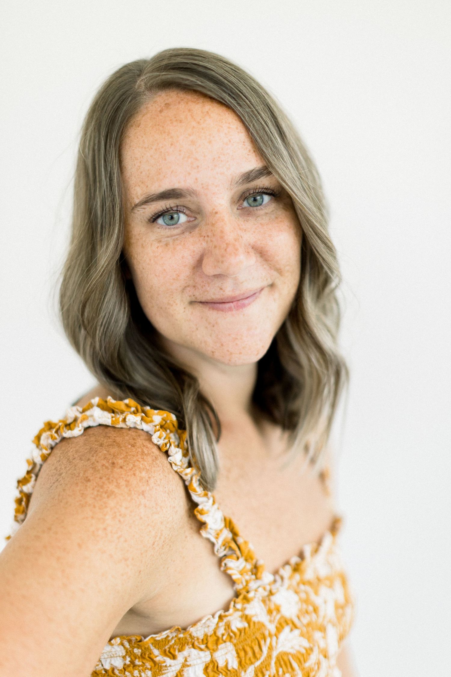Young woman with freckles and gray-blonde wavy hair smiles softly, wearing a gold floral top against white background.