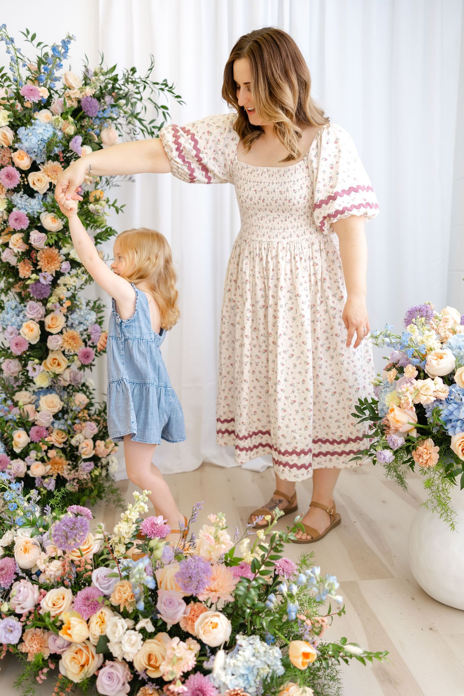Mother and daughter in floral dresses dancing among colorful spring flower arrangements indoors.