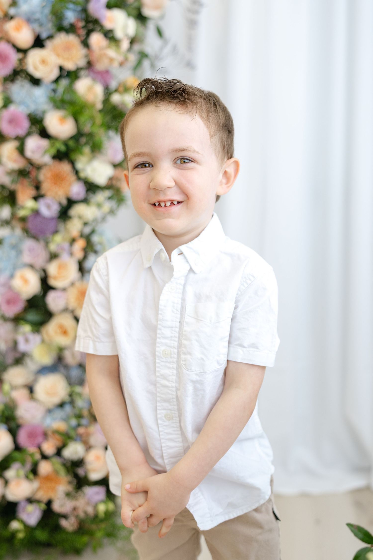 Smiling young boy in white shirt poses in front of colorful floral backdrop with pink and peach roses.