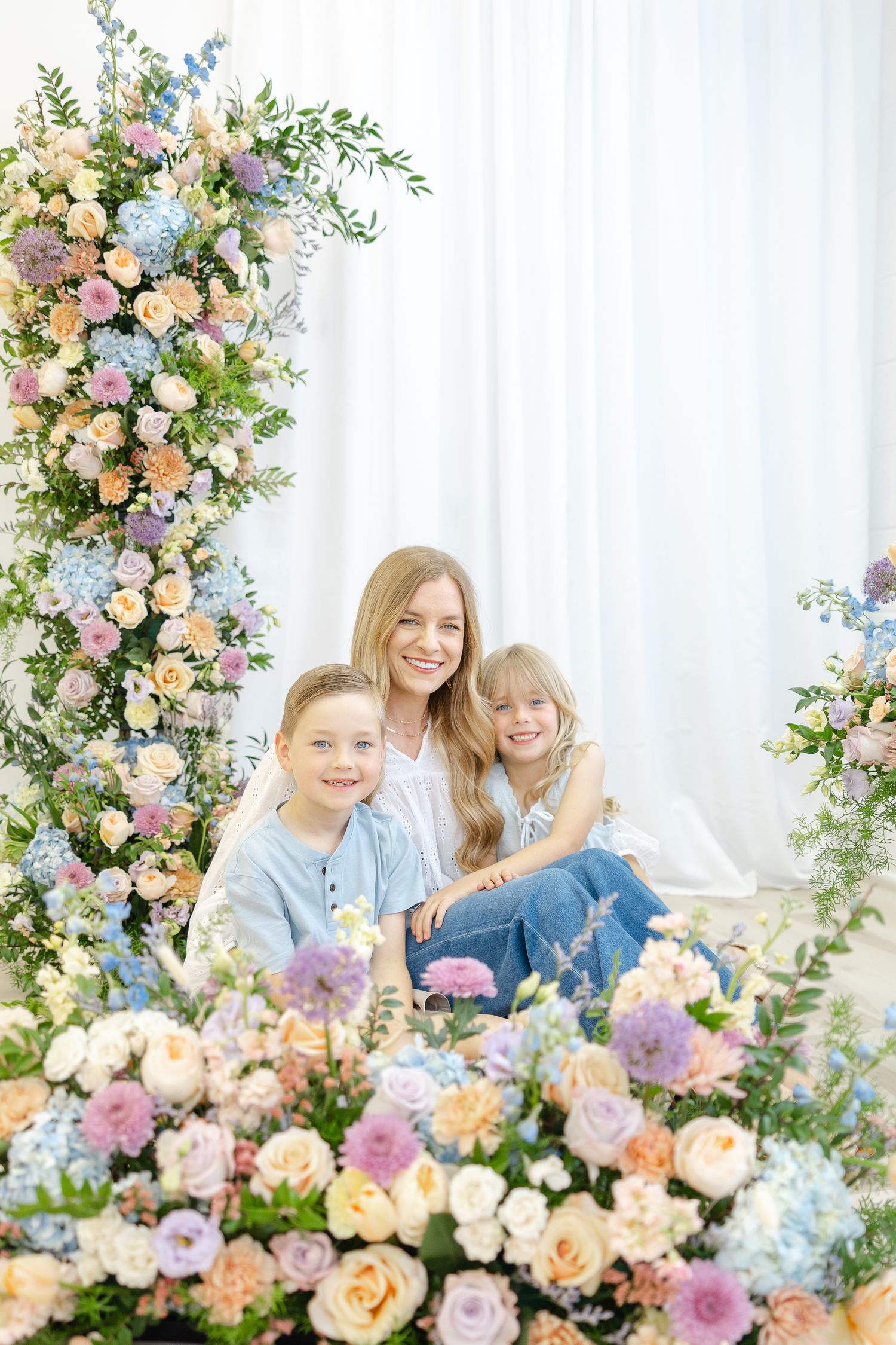 Woman and two children smiling among pastel floral arrangements in a bright studio setting.