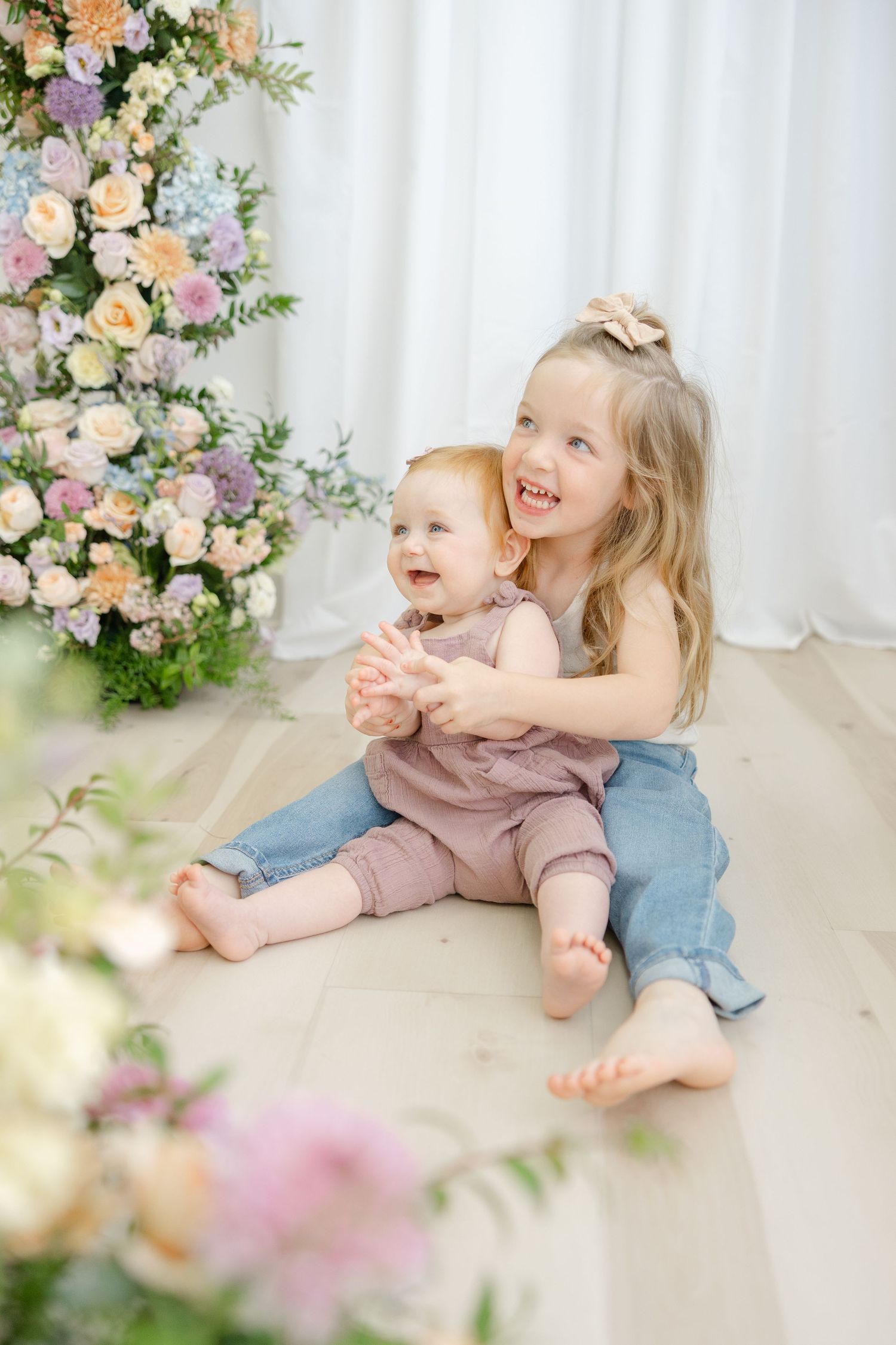 Two happy sisters sitting on floor surrounded by pastel floral arrangements in a bright studio setting.