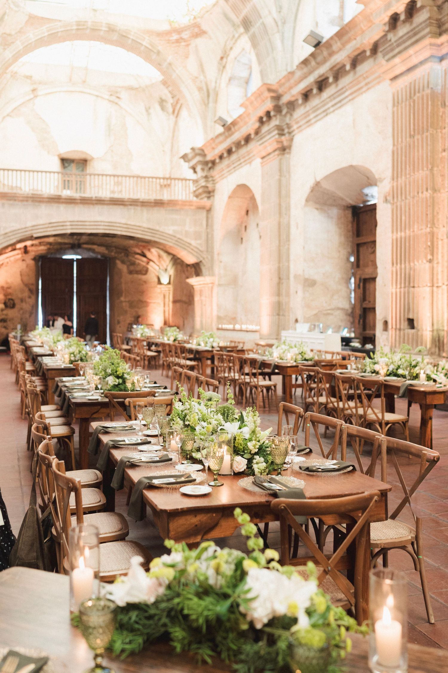 Elegant wedding reception in a historic stone venue with long wooden tables, greenery centerpieces, and cross-back chairs.